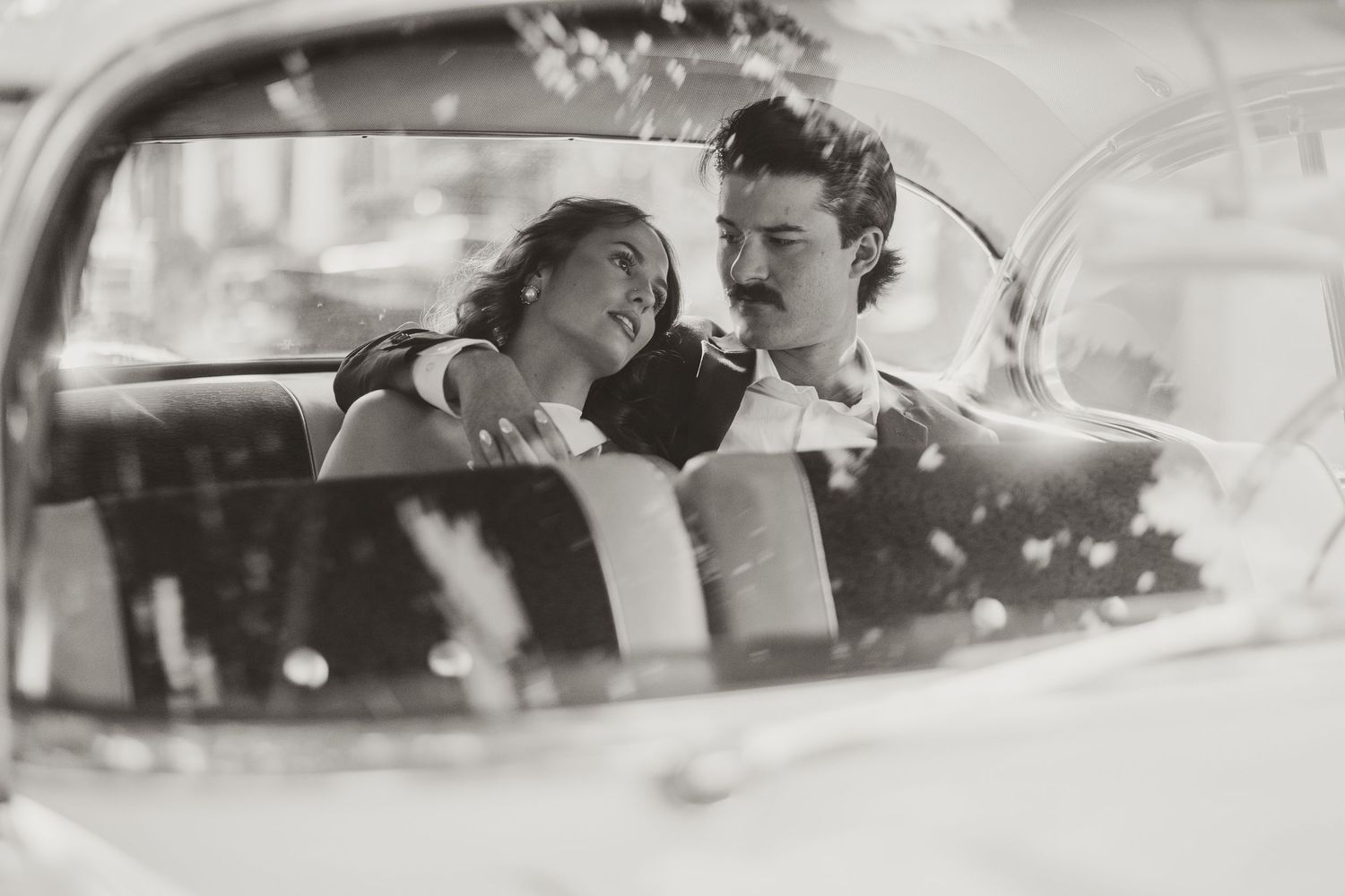 Black and white photo of a smiling couple sharing an intimate moment in the front seat of a vintage car.