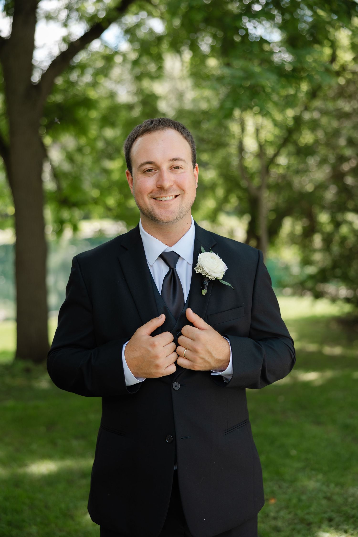 Smiling groom in black suit with white boutonniere adjusting tie outdoors in green park setting.