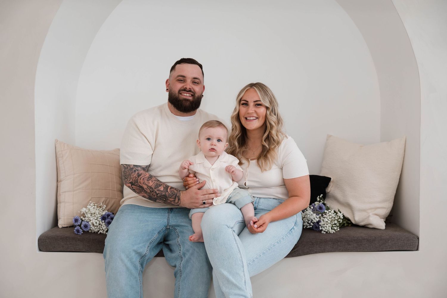 Family portrait of parents and baby sitting together on a bench in a bright minimalist studio setting.