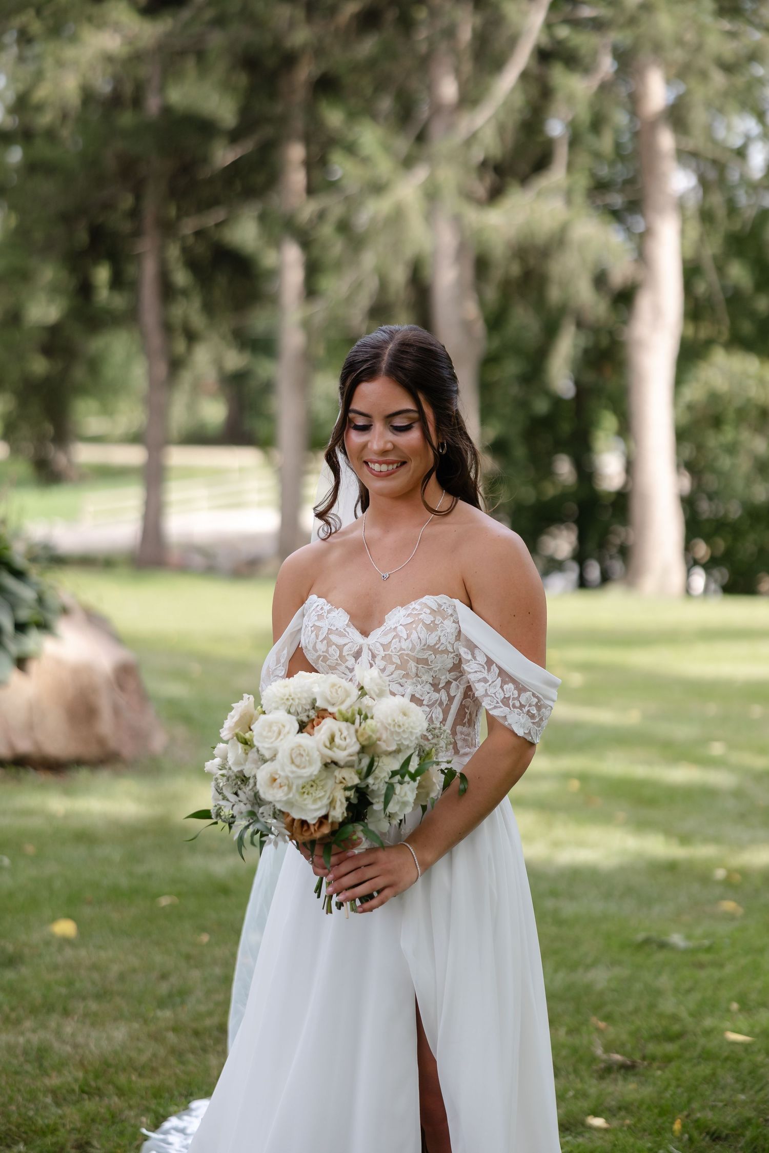 Smiling bride in an off-the-shoulder lace wedding gown holding a white rose bouquet on a sunlit green lawn with tall trees.