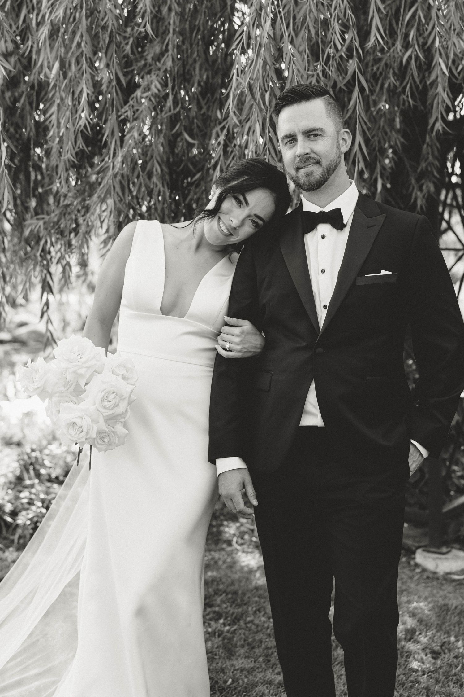 Black and white photo of bride and groom embracing under a weeping willow tree.