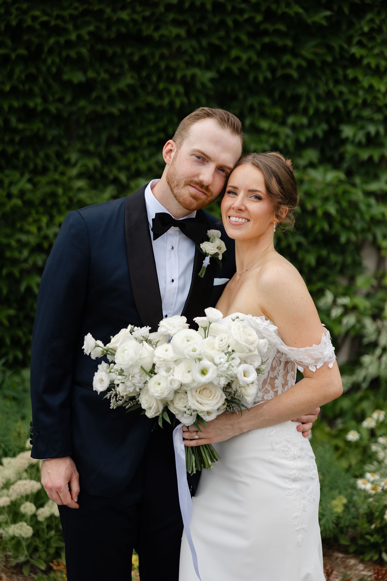 A bride and groom pose together in a garden, she holds a white floral bouquet, wearing an off-shoulder gown, he wears a black tuxedo.