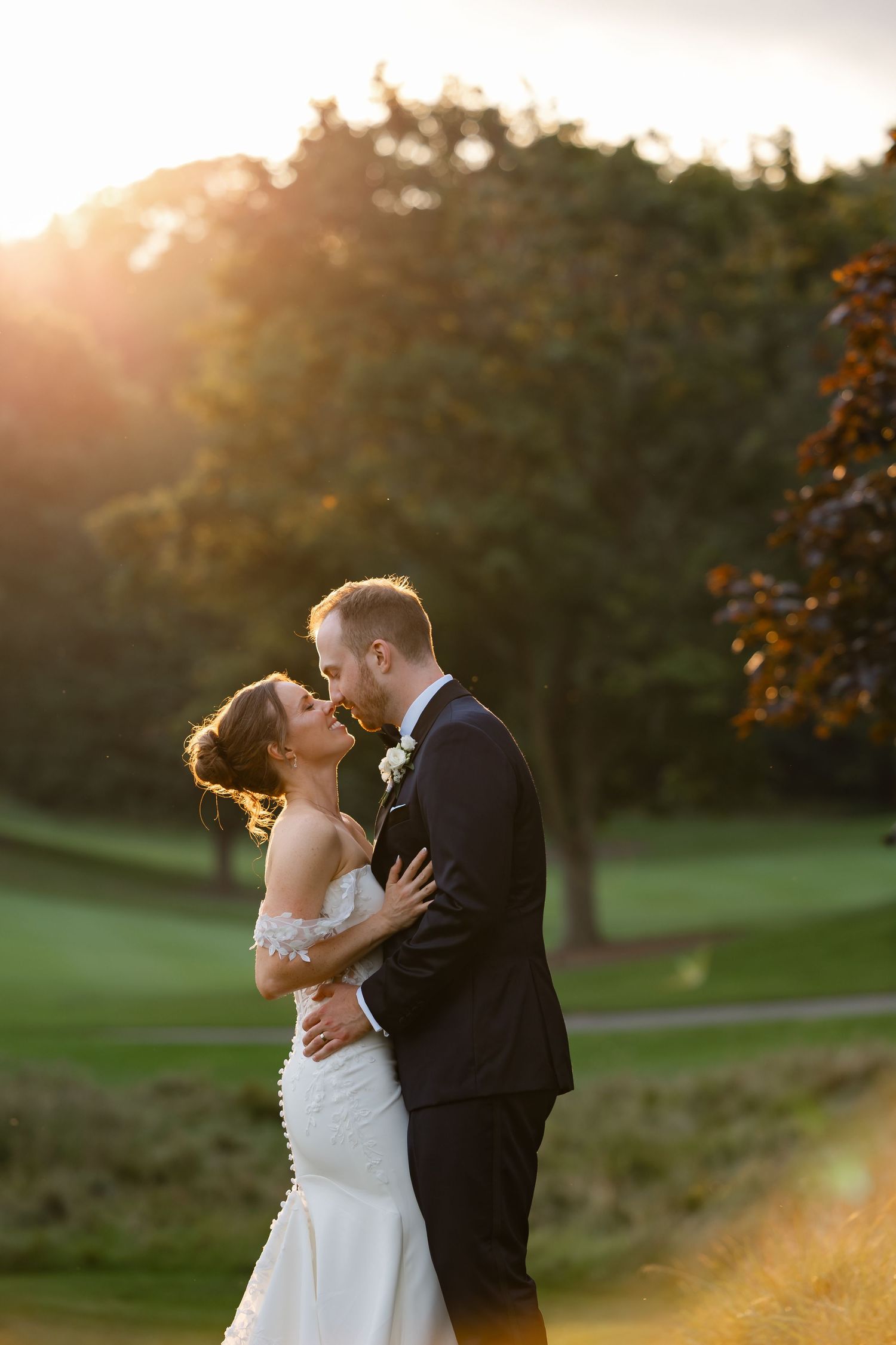 Bride and groom share a romantic golden hour kiss outdoors, surrounded by lush green trees at their wedding.