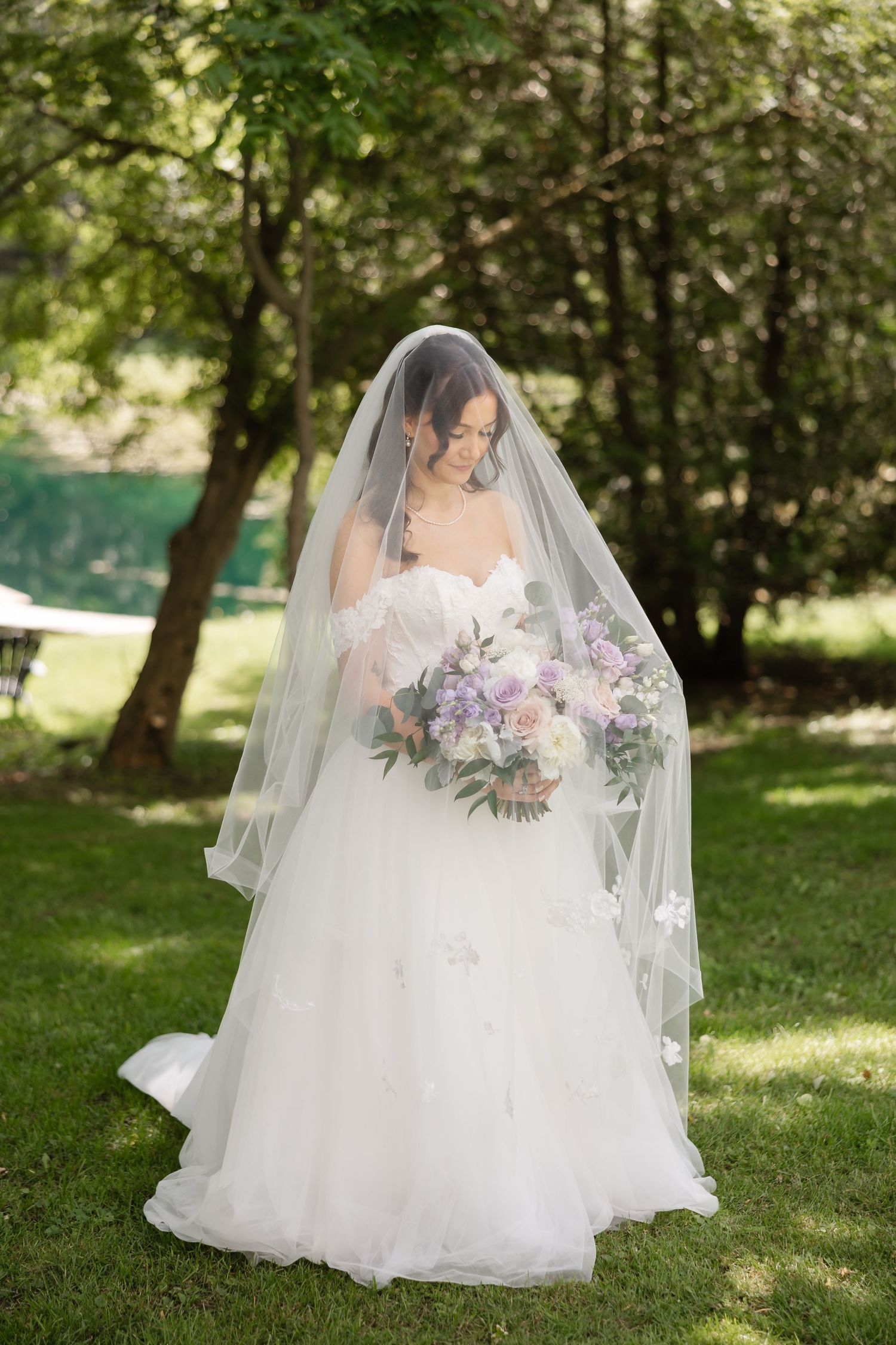 Elegant bride in strapless ballgown holding pastel bouquet with veil draped over face outdoors.