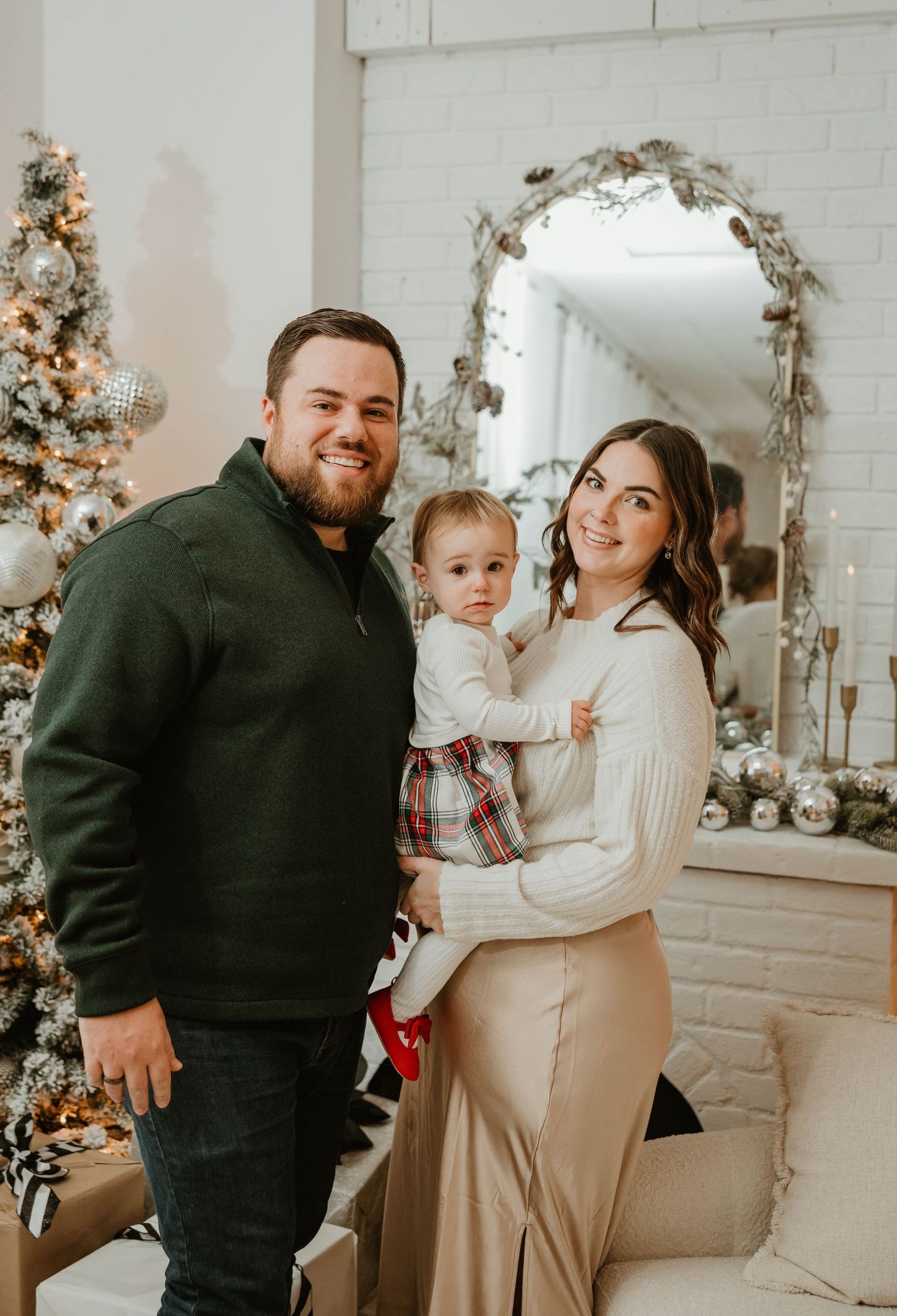 Family of three posing by decorated Christmas tree, mother holding toddler in plaid outfit.