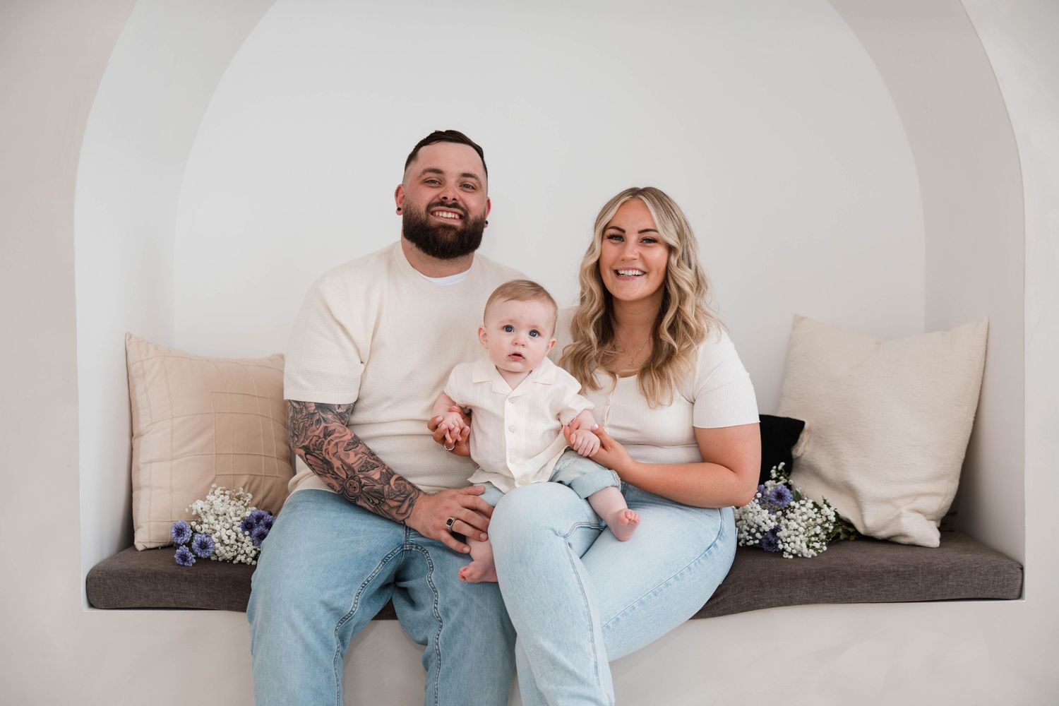 Family portrait of tattooed father, blonde mother, and baby sitting in a white arched alcove bench.