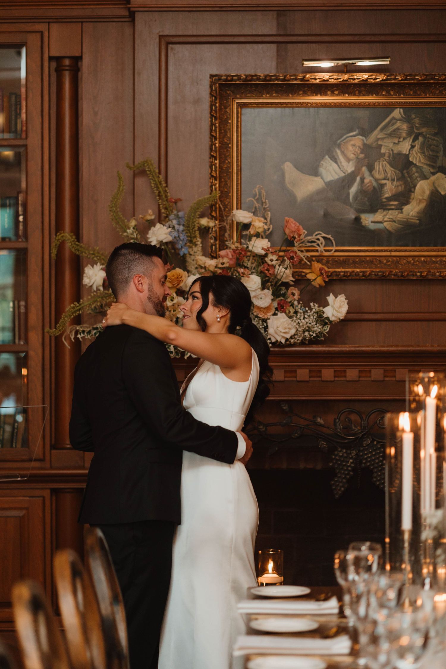 Bride and groom sharing a romantic dance near candlelit table in elegant wood-paneled room.