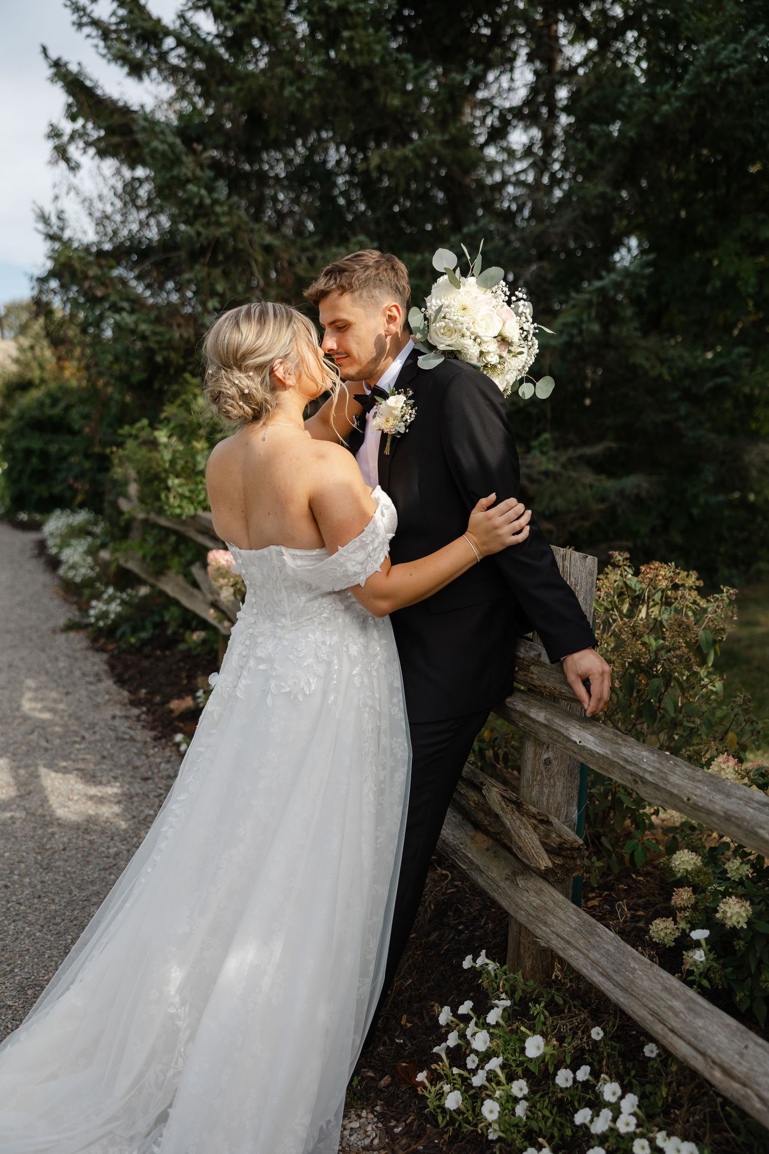 Bride in an off-shoulder lace wedding gown and groom in a black tuxedo share an intimate moment beside a rustic wooden fence.