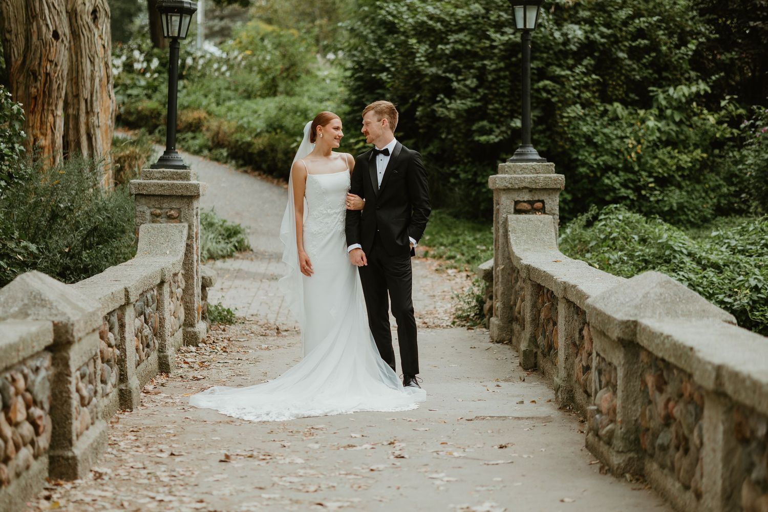 Bride and groom pose on a stone bridge path surrounded by lush greenery, she in a flowing white gown with train.
