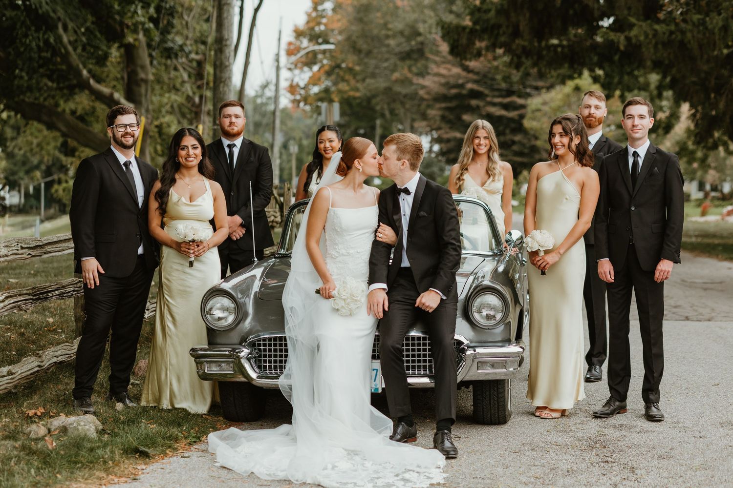 Wedding party posing with vintage classic car, bridesmaids in gold gowns and groomsmen in black suits.