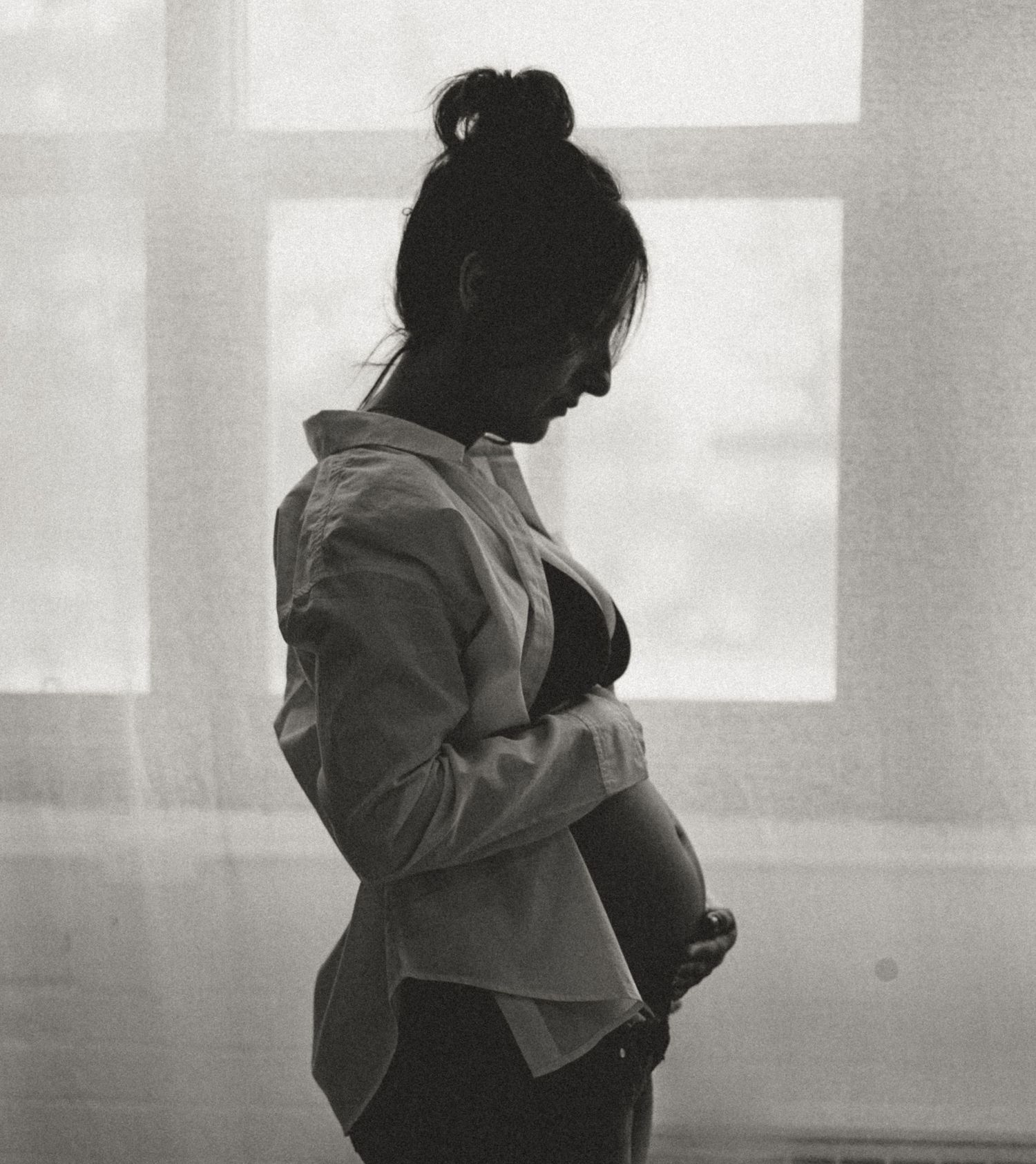 Black and white silhouette of a pregnant woman cradling her baby bump by a bright window.