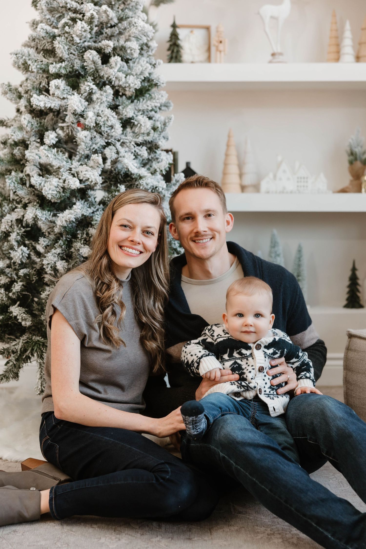 Smiling couple with baby in festive sweater sitting by flocked Christmas tree indoors.