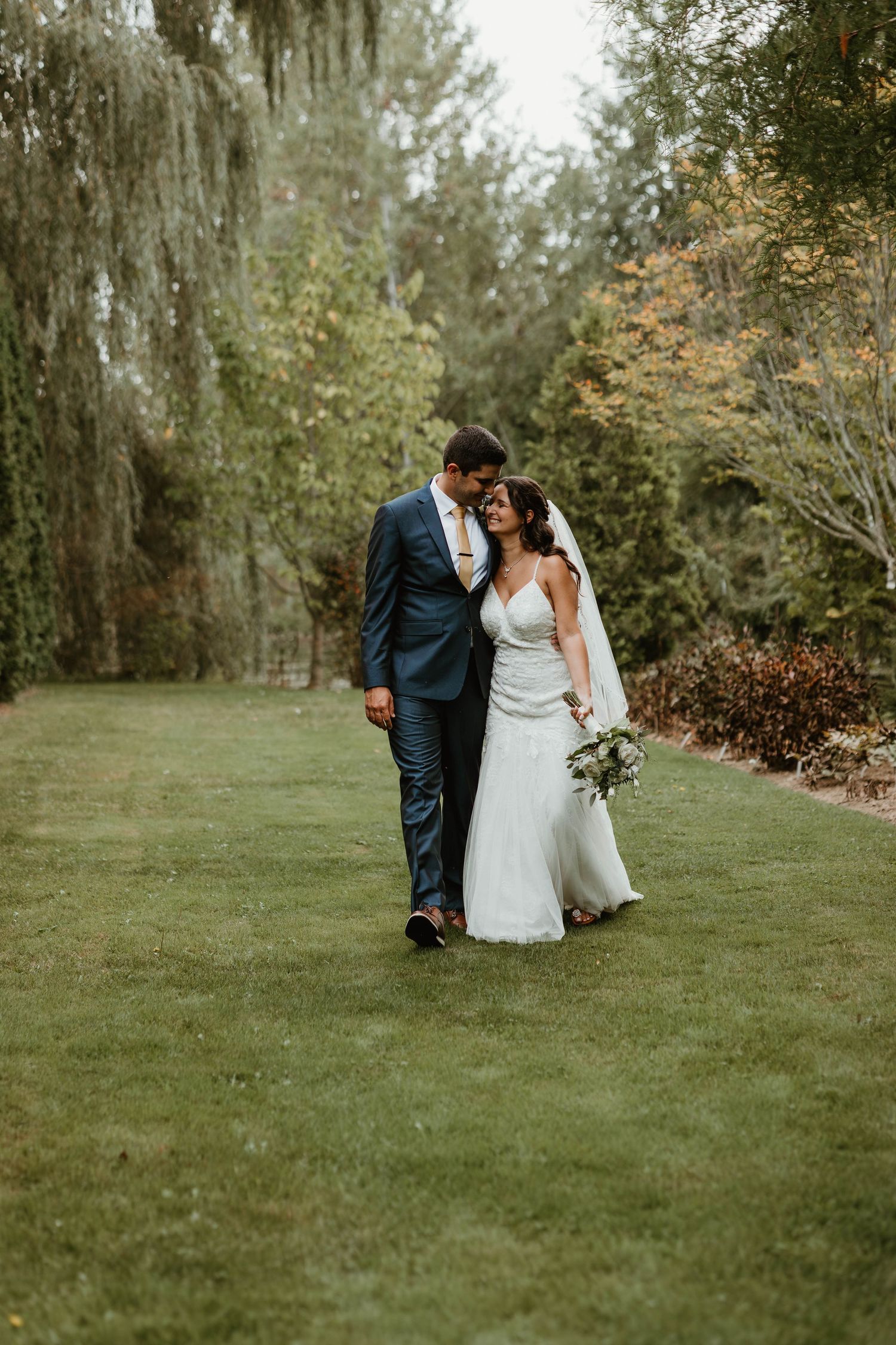 Bride and groom walk closely together on lush green lawn surrounded by weeping willow trees.