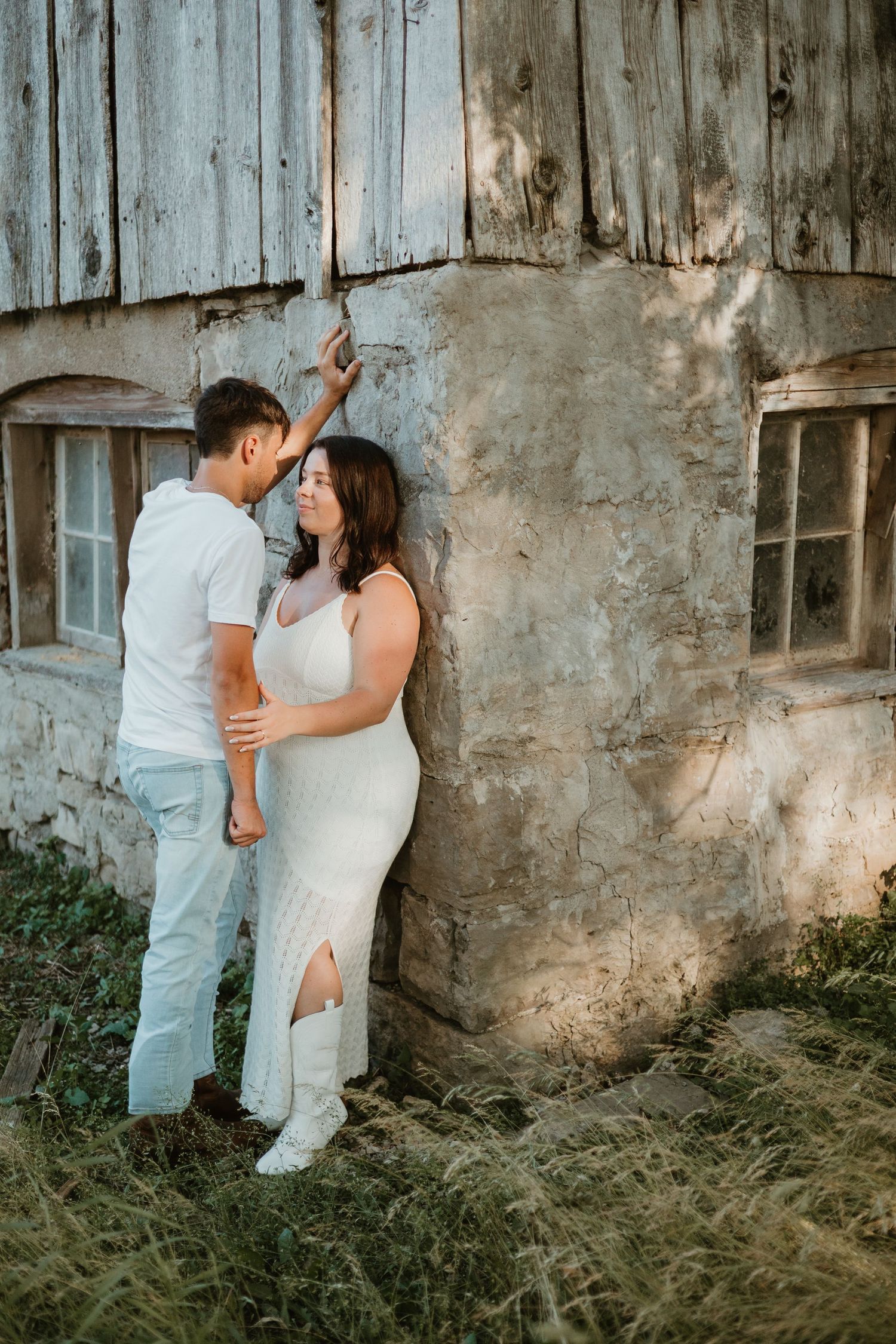 Couple in white outfits share a romantic moment leaning against a rustic weathered barn wall outdoors.