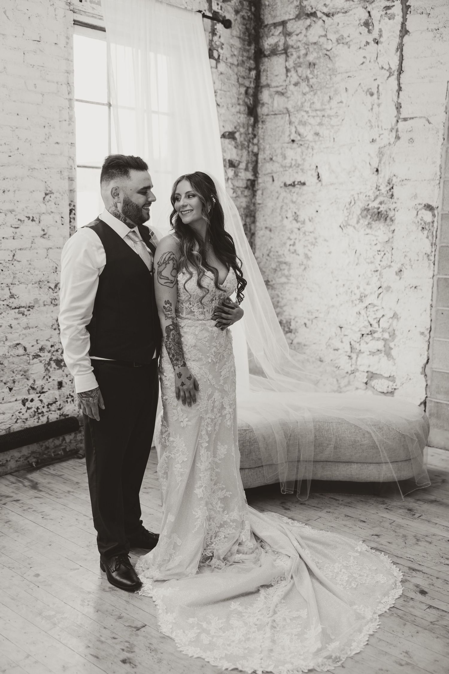 Black and white portrait of tattooed groom and bride in lace gown with cathedral veil in rustic brick venue.