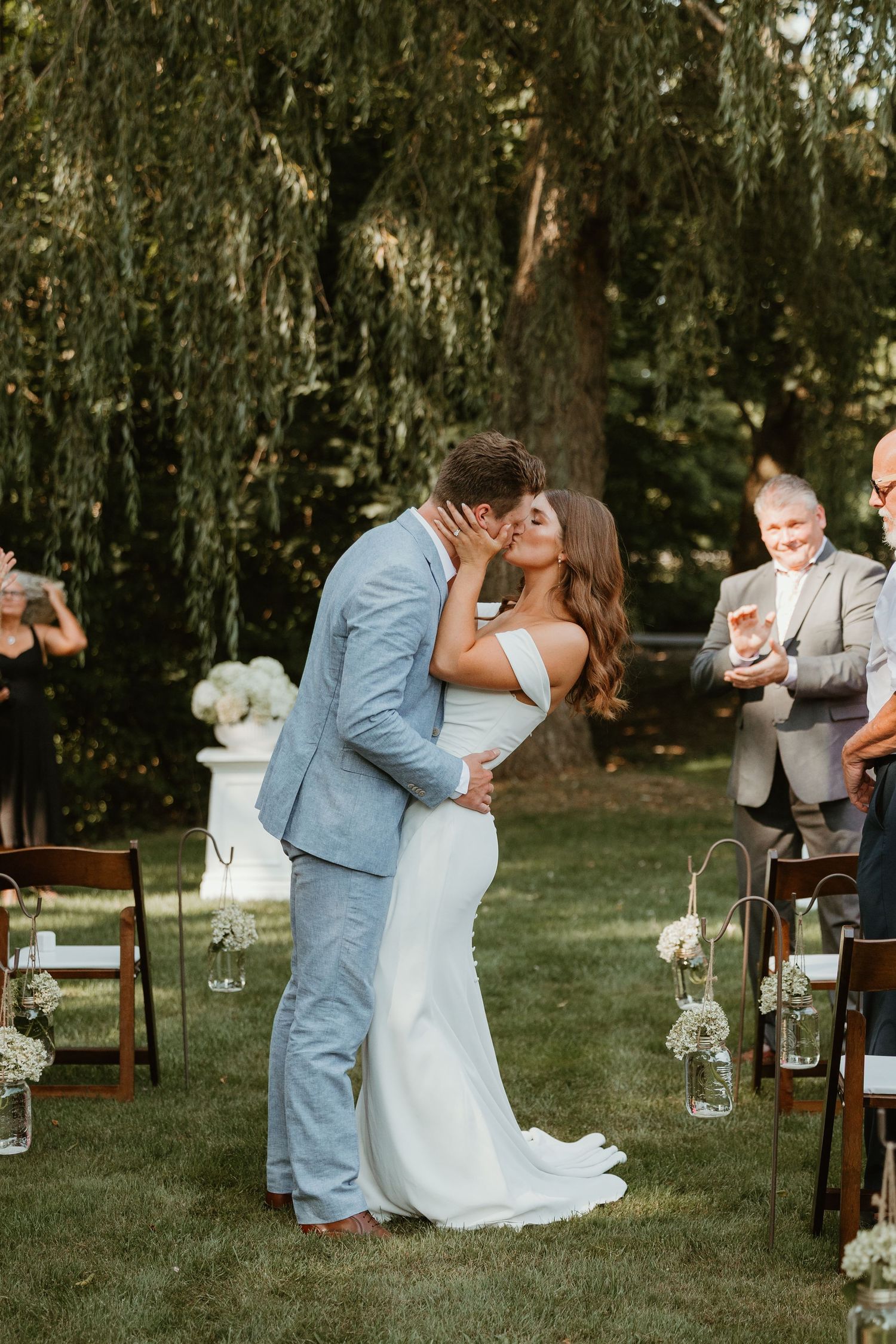 Bride and groom sharing first kiss at outdoor garden wedding ceremony under willow tree.