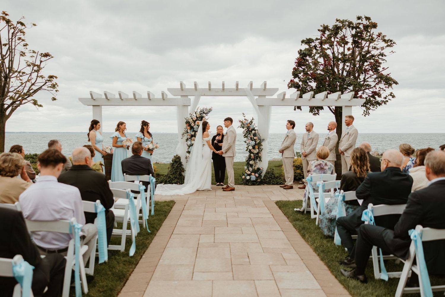 Outdoor wedding ceremony under white pergola with ocean view, bride and groom at altar surrounded by wedding party.