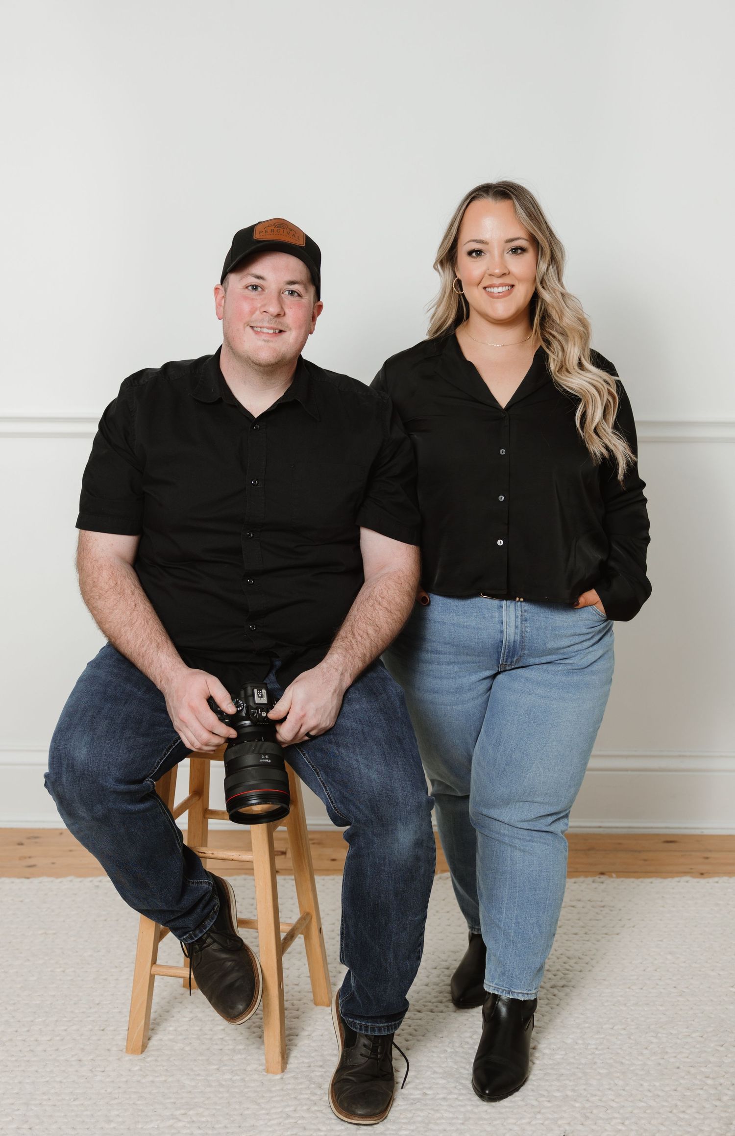 A man holding a camera and a woman in black outfits pose together for a professional photography studio portrait.