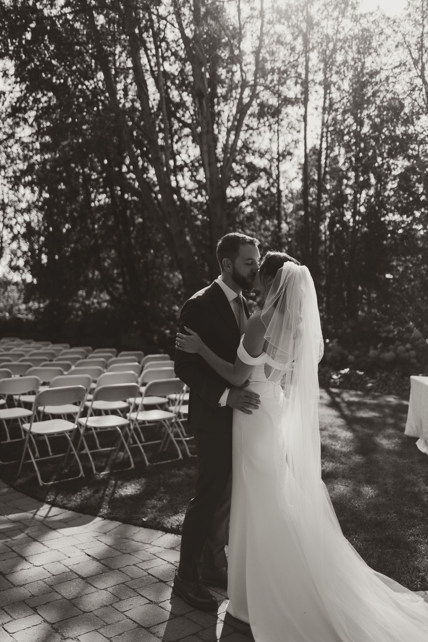 Black and white photo of newlyweds kissing at outdoor ceremony venue surrounded by trees.