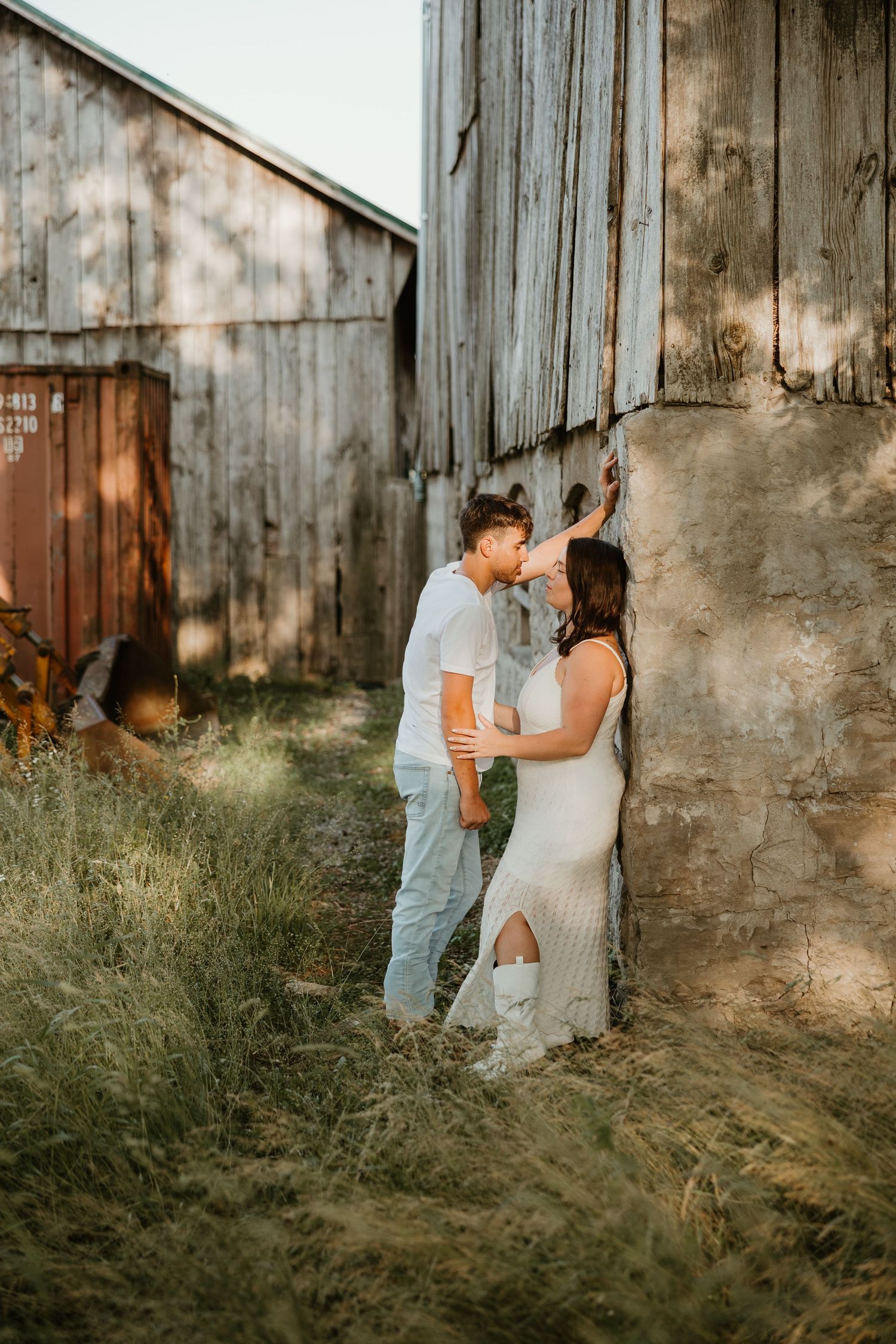 Couple sharing a kiss against a rustic barn wall surrounded by tall grass in golden light.