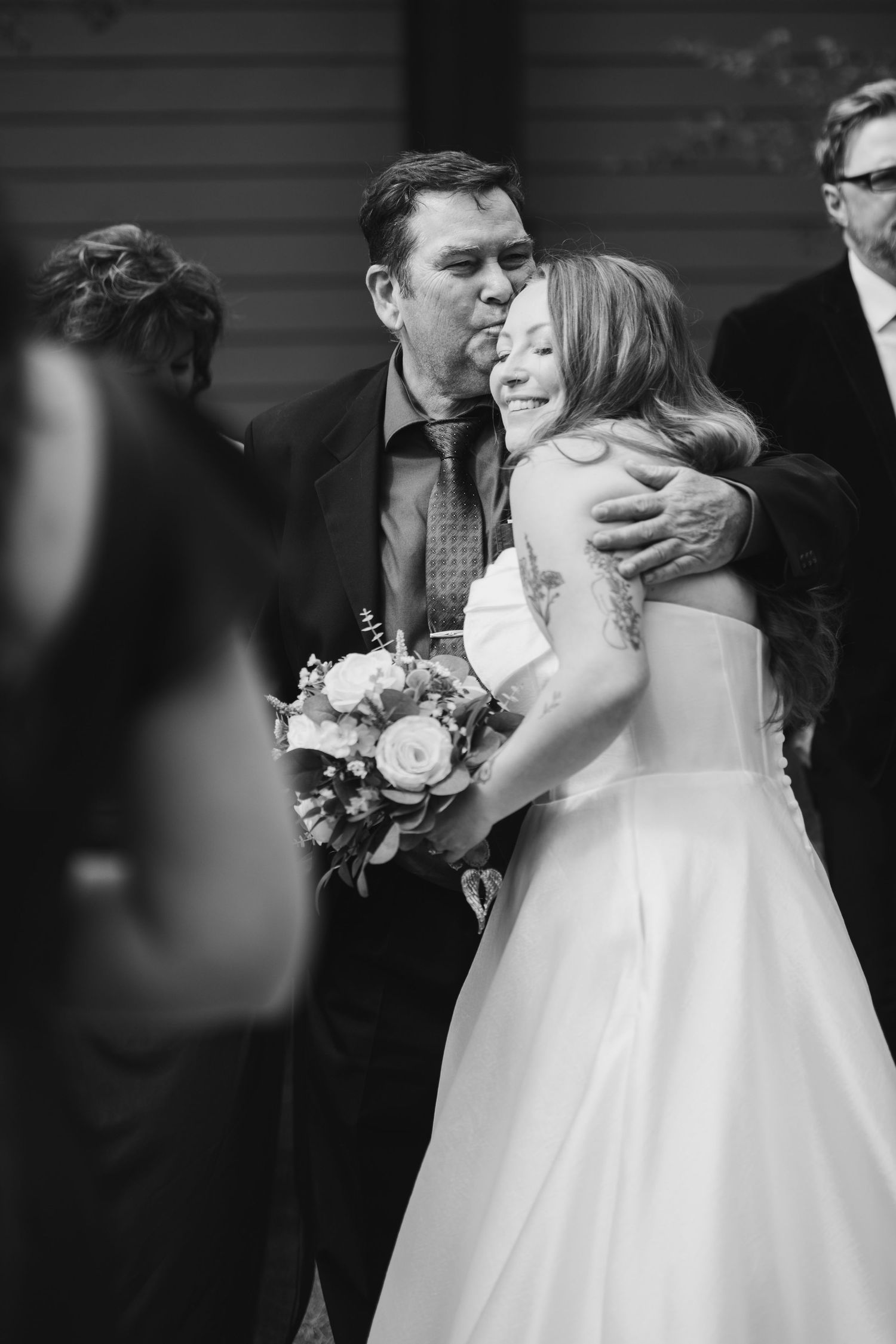 Bride holding floral bouquet shares a warm embrace with a guest in black and white wedding photo.