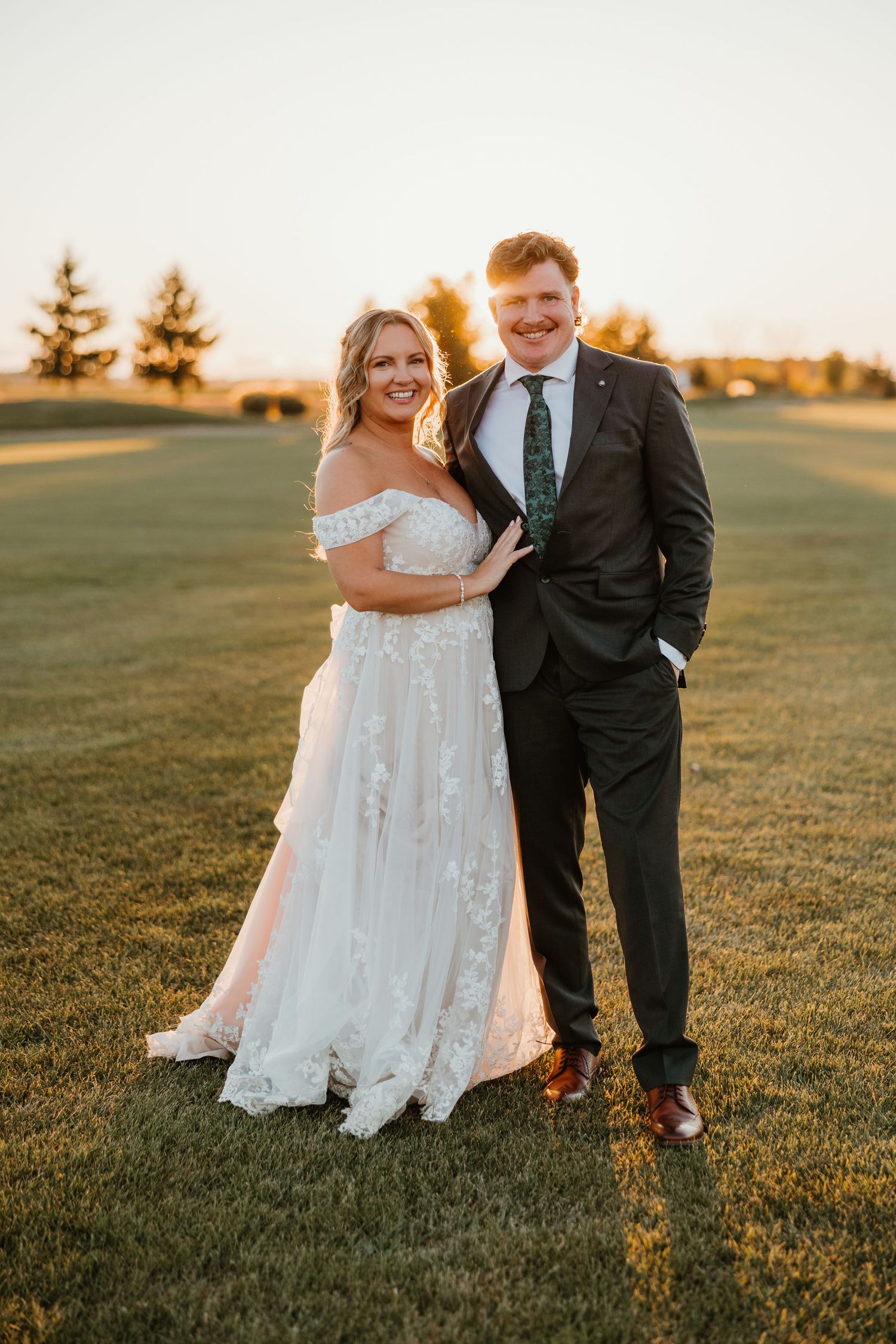Bride in off-shoulder floral gown and groom in dark suit posing at golden hour on green lawn.