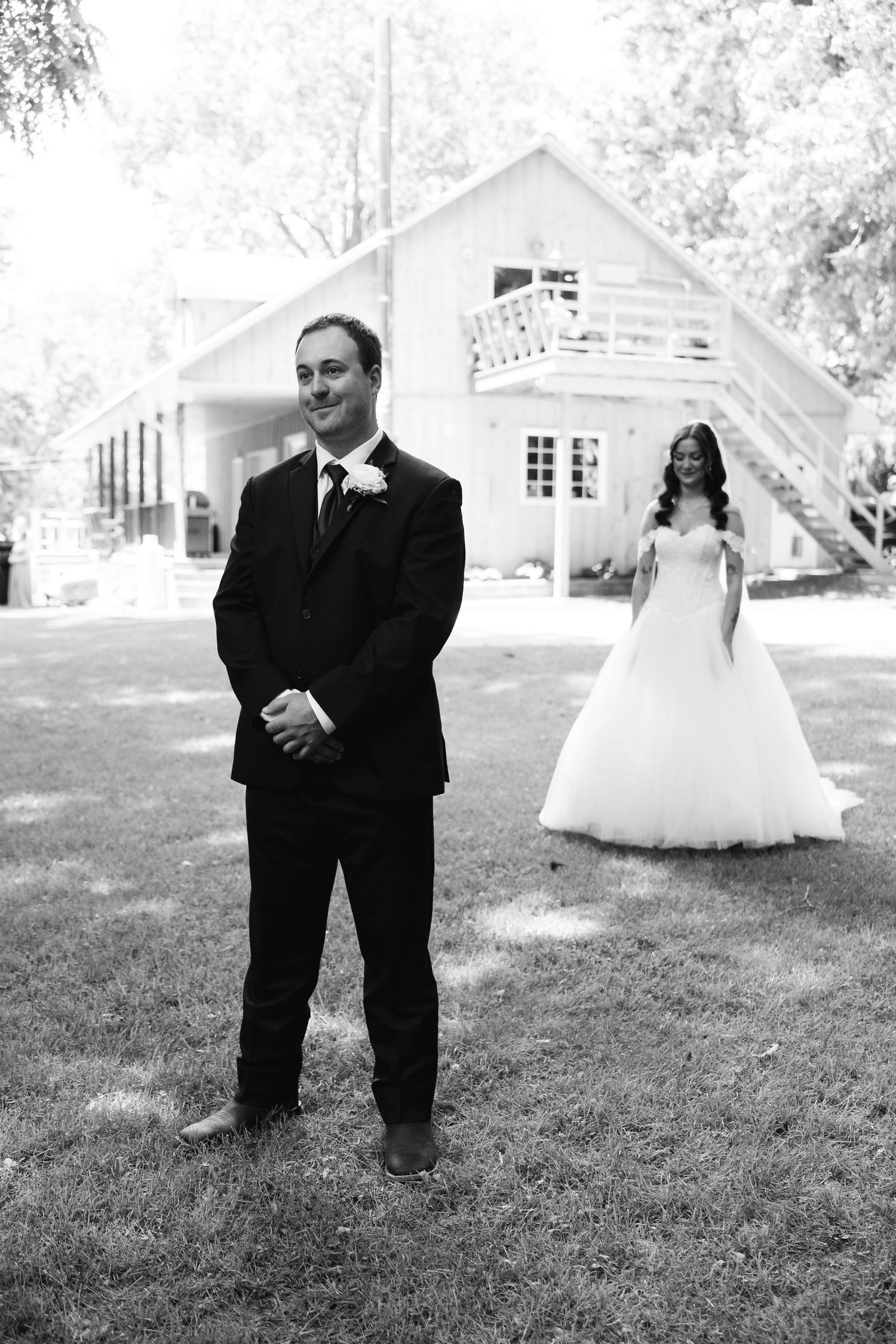 Black and white photo of groom smiling as bride approaches in white gown outside a white barn venue.