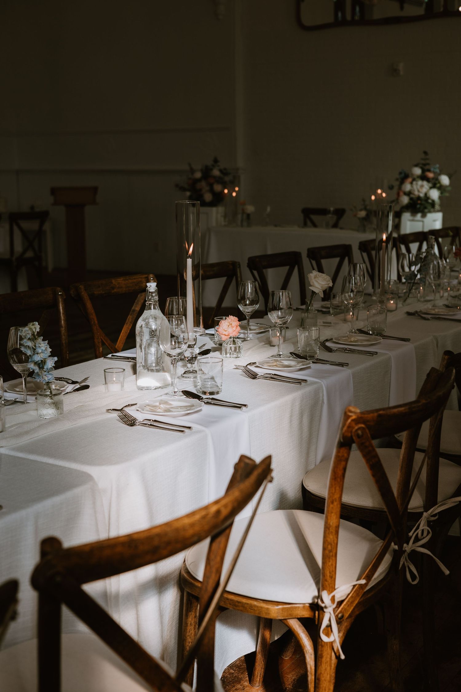 Elegant wedding reception tables with white linens, wooden cross-back chairs, candles, and floral centerpieces.