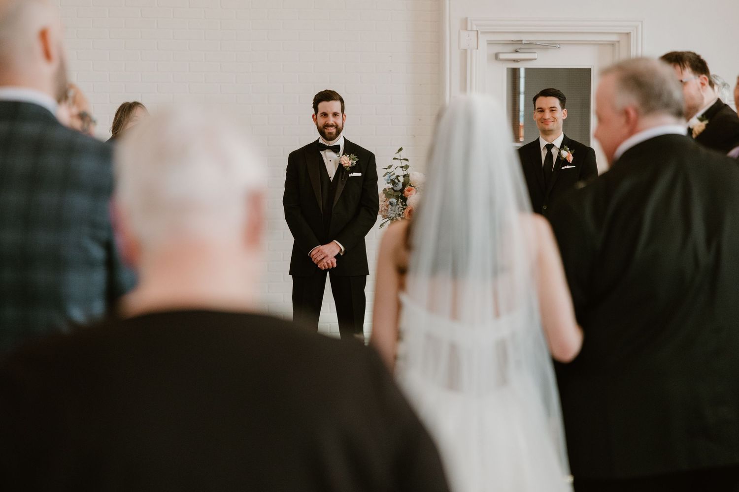 Groom watches bride walk down the aisle during elegant indoor wedding ceremony.