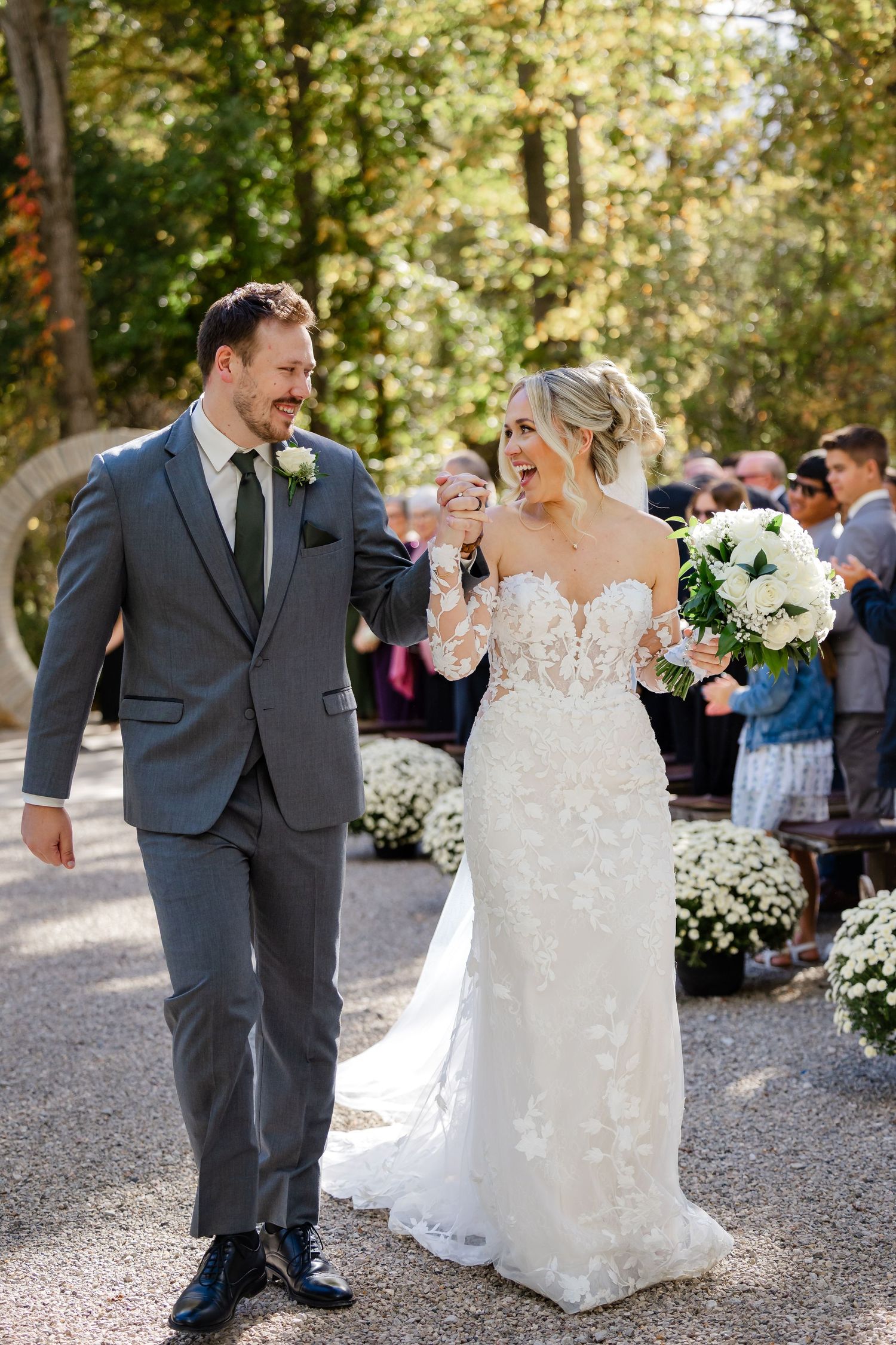 Smiling bride in a lace wedding gown and groom in a gray suit walk hand in hand down an outdoor aisle holding a white floral bouquet.