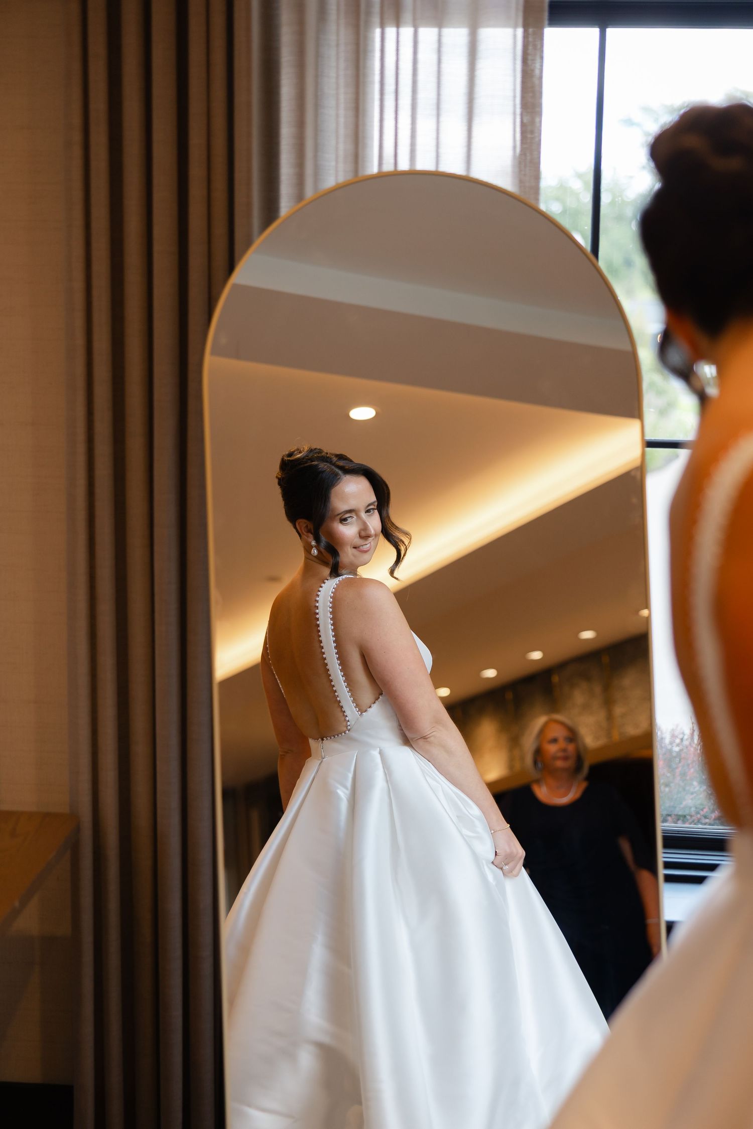 Bride in a white satin ball gown with pearl-trimmed open back looks over her shoulder at her reflection in an arched mirror.