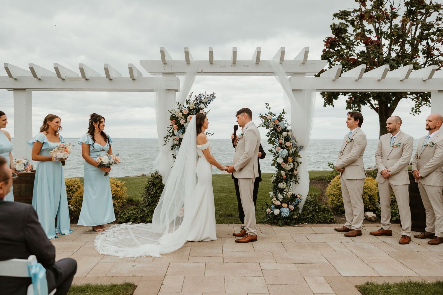 Outdoor oceanview wedding ceremony under white pergola with blue bridesmaid dresses.
