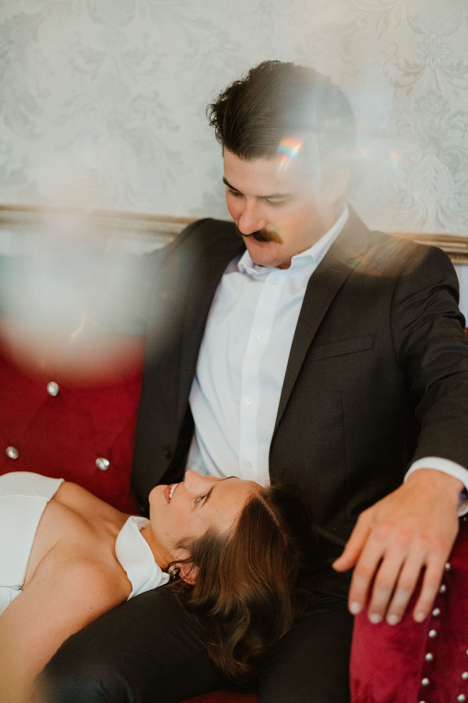 Groom in black suit gazes at bride lying across his lap on a red velvet sofa in an elegant room.