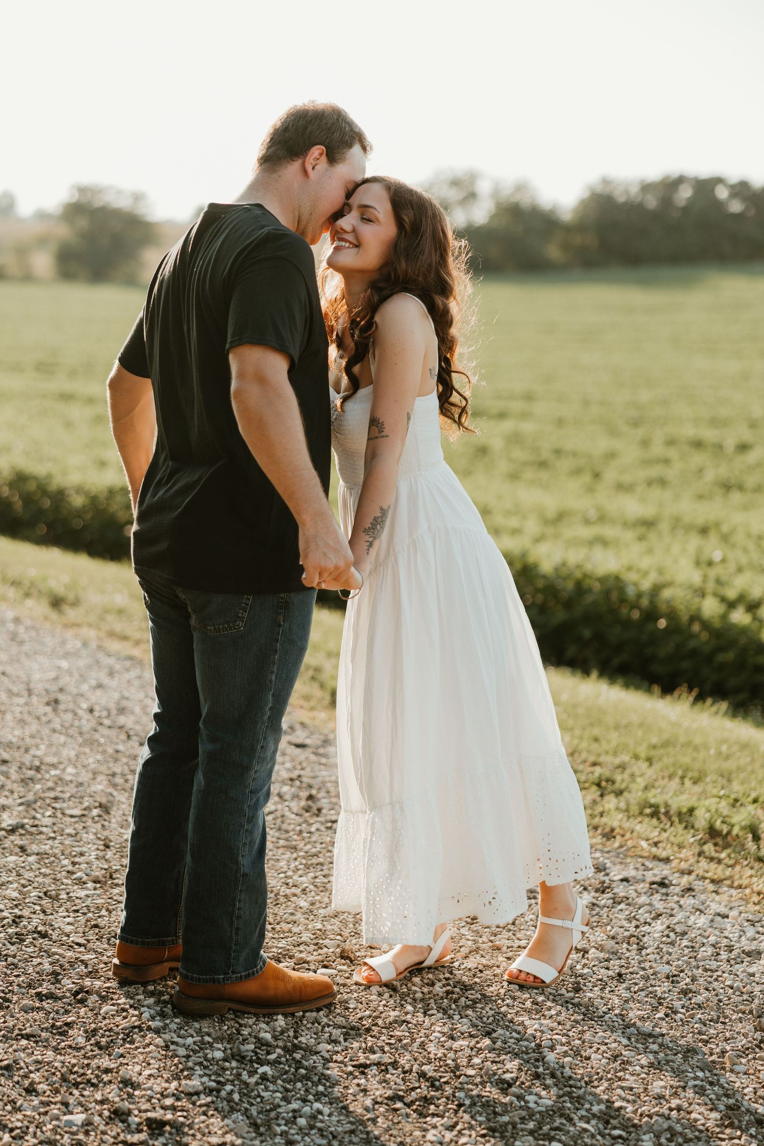 Laughing couple holding hands on a sunlit gravel path surrounded by green fields at sunset.