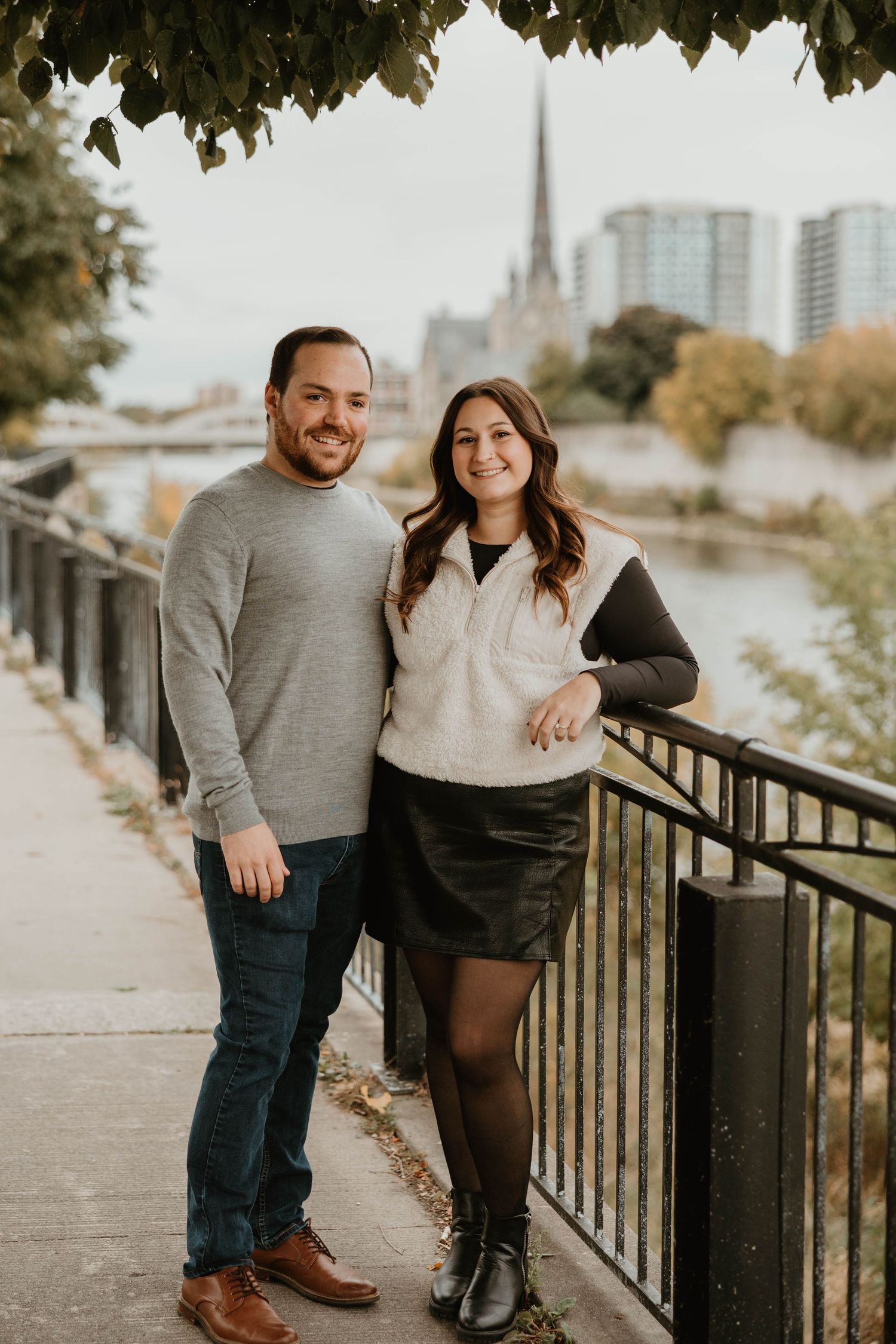 Smiling couple posing by riverside fence with church steeple and city behind them.