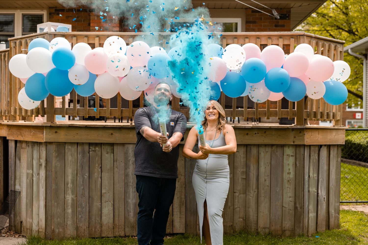 Couple at gender reveal party with blue confetti explosion in front of balloon arch.