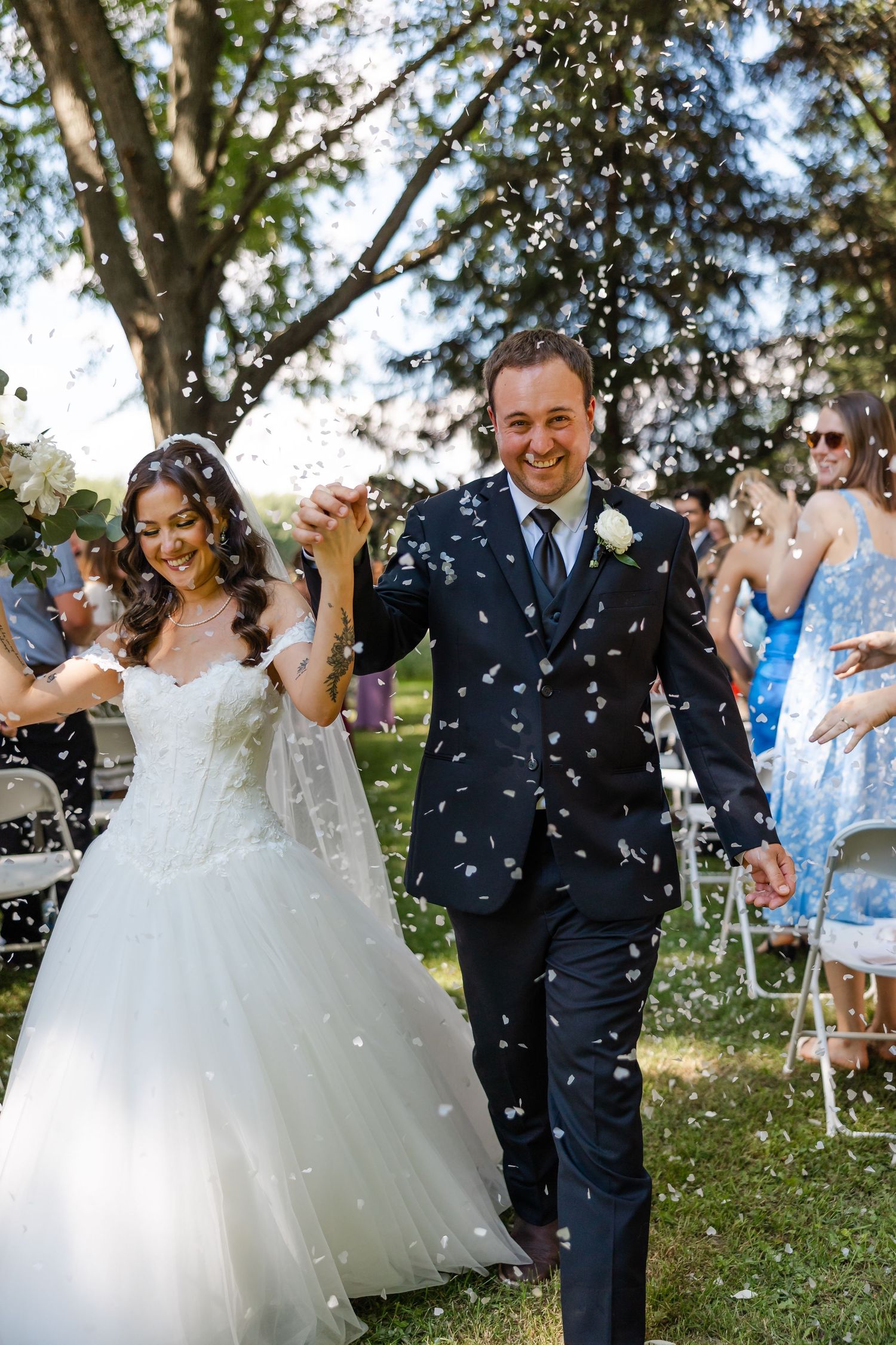 Joyful newlyweds walk down outdoor aisle as guests toss white confetti petals in sunny garden wedding ceremony.