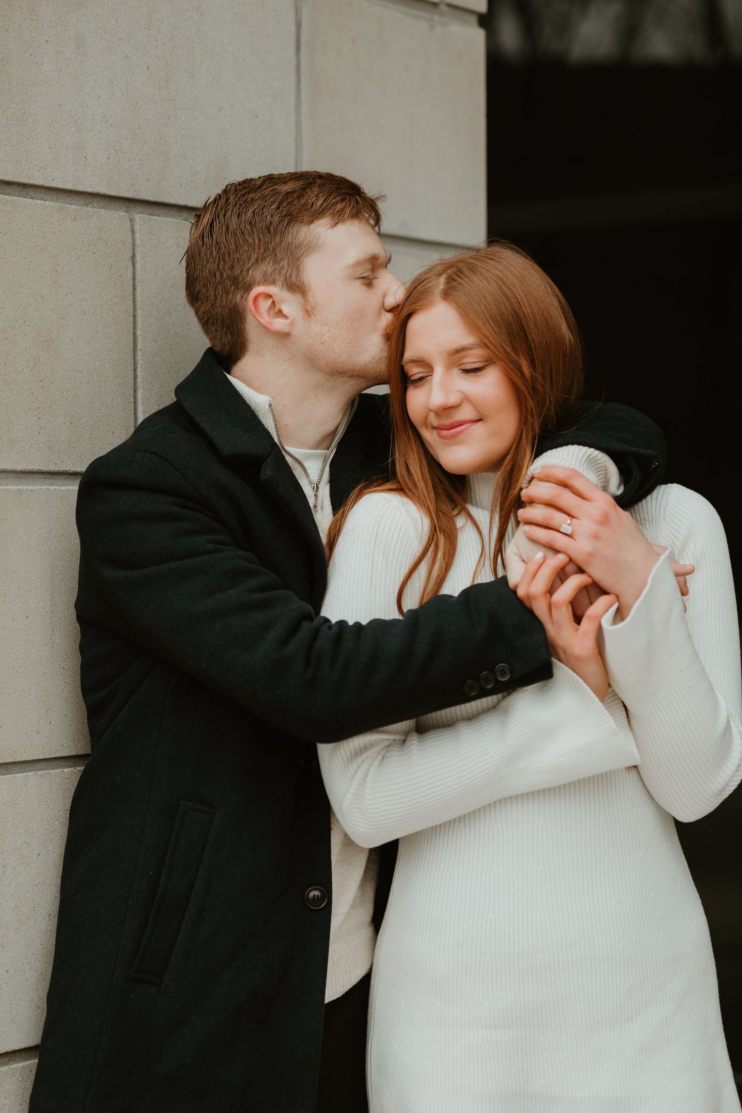 A young man in a black coat kisses a redheaded woman in a white dress against a stone wall.