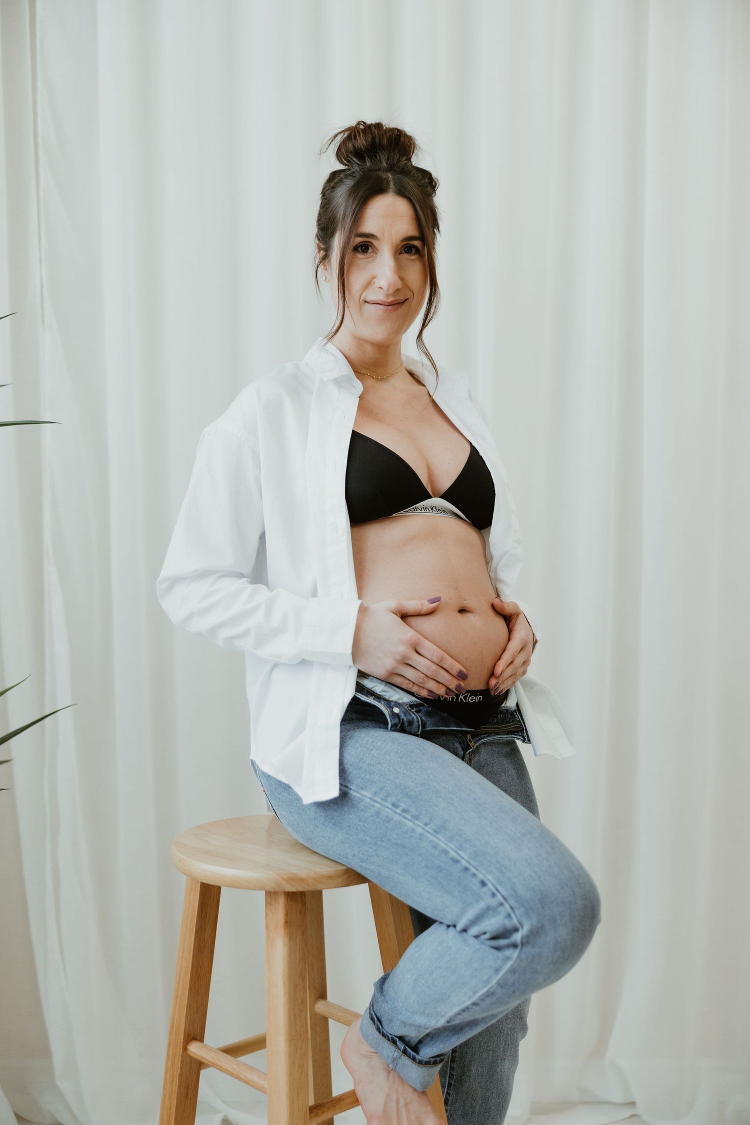 Smiling pregnant woman in black bra and white shirt sitting on a stool holding her baby bump.