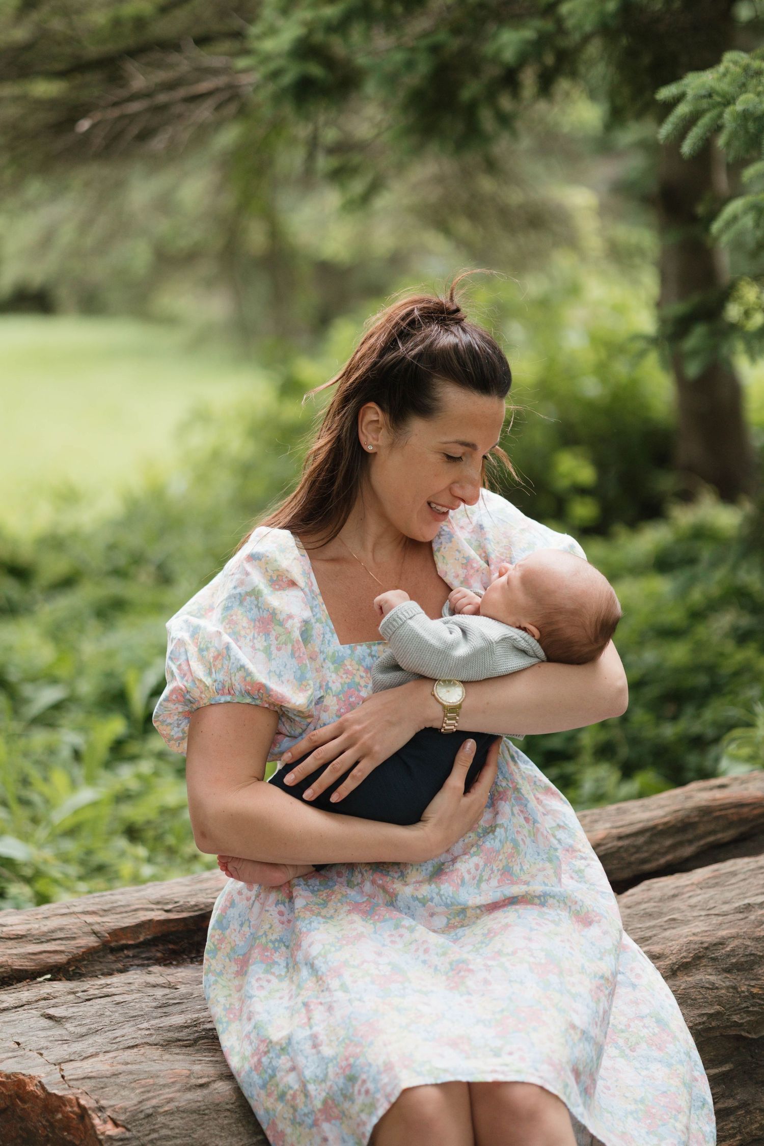 Mother in floral dress sitting on a log cradling her newborn baby in a lush green park.