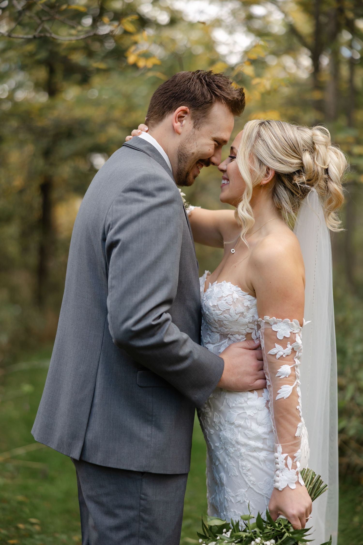 Bride in a lace gown with a veil and groom in a gray suit touch noses in a loving embrace among autumn trees at an outdoor wedding.