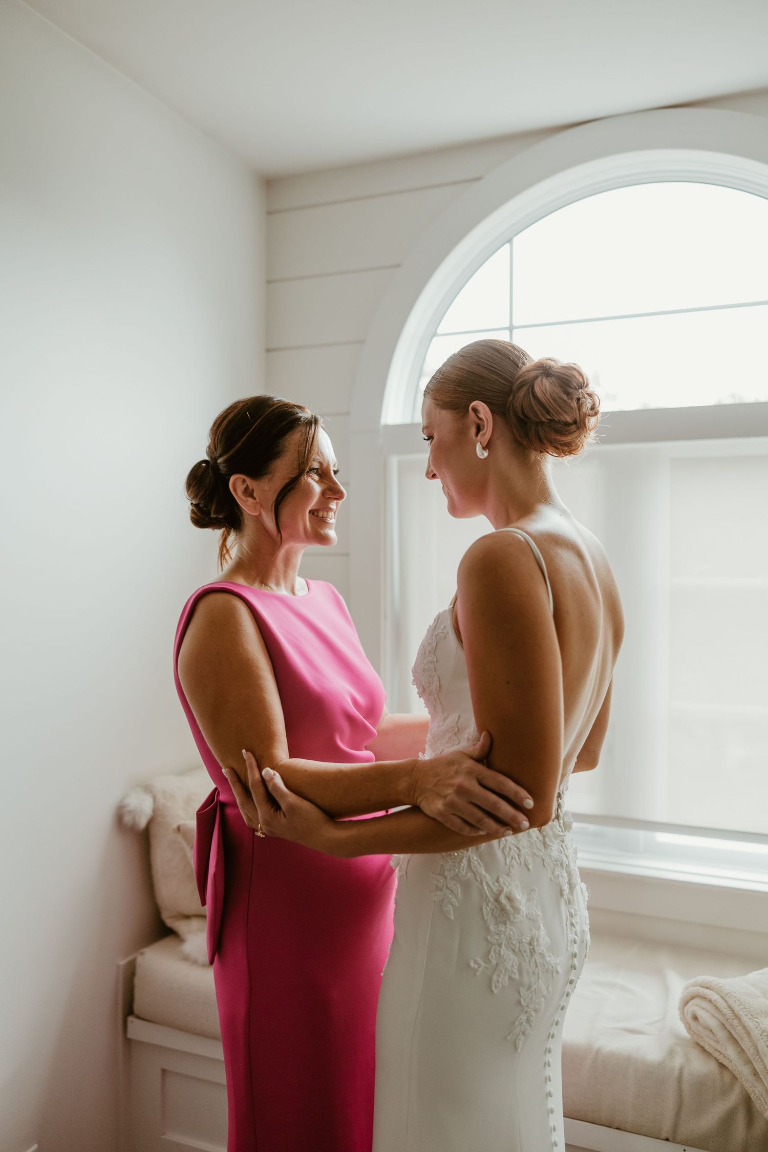 Bride in lace gown shares a heartfelt moment with her mother in pink dress by an arched window.