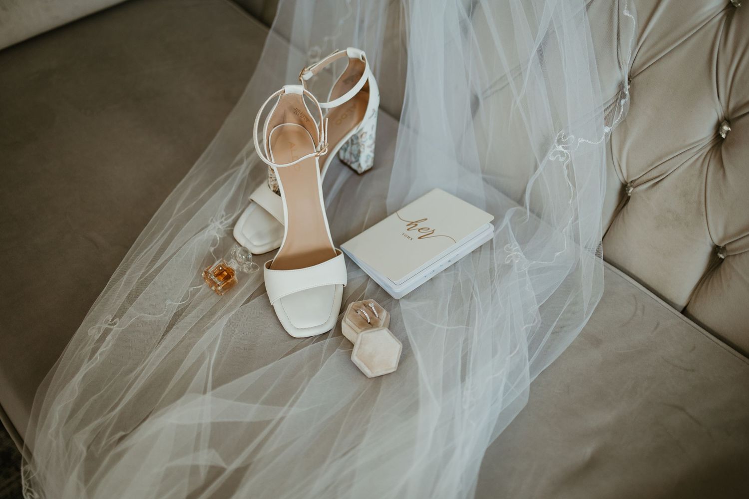 White block-heel bridal sandals, veil, perfume bottles, and monogrammed book on velvet sofa.