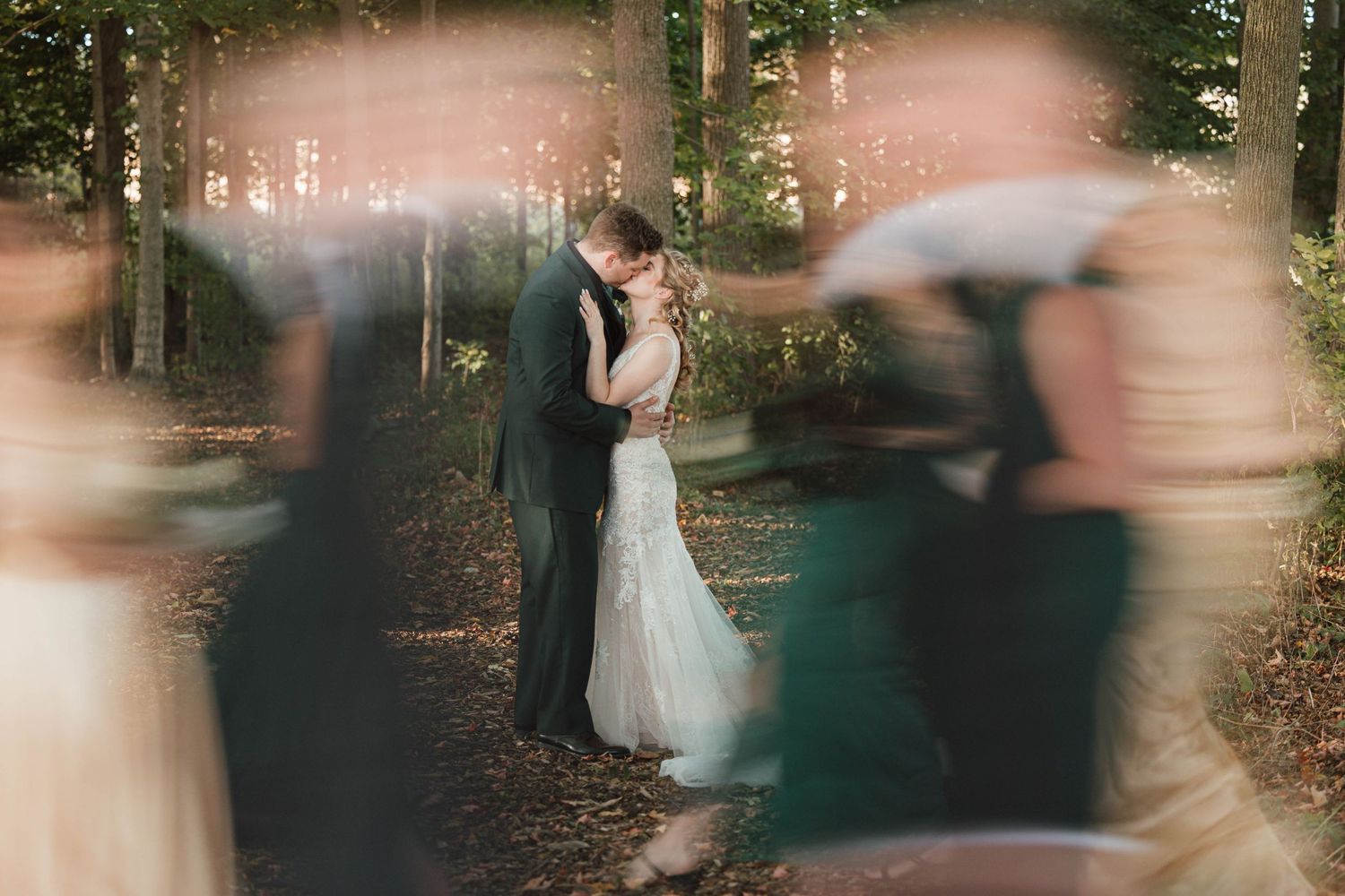 Couple shares romantic kiss in sunlit forest surrounded by blurred wedding guests in dreamy bokeh photography.