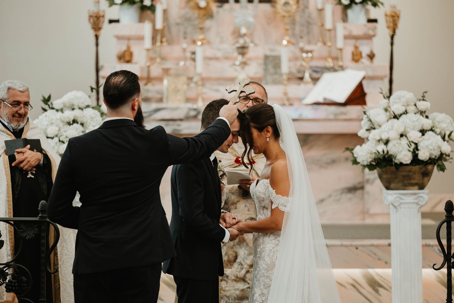 Groom places wedding crown on bride during traditional church ceremony with white floral arrangements.