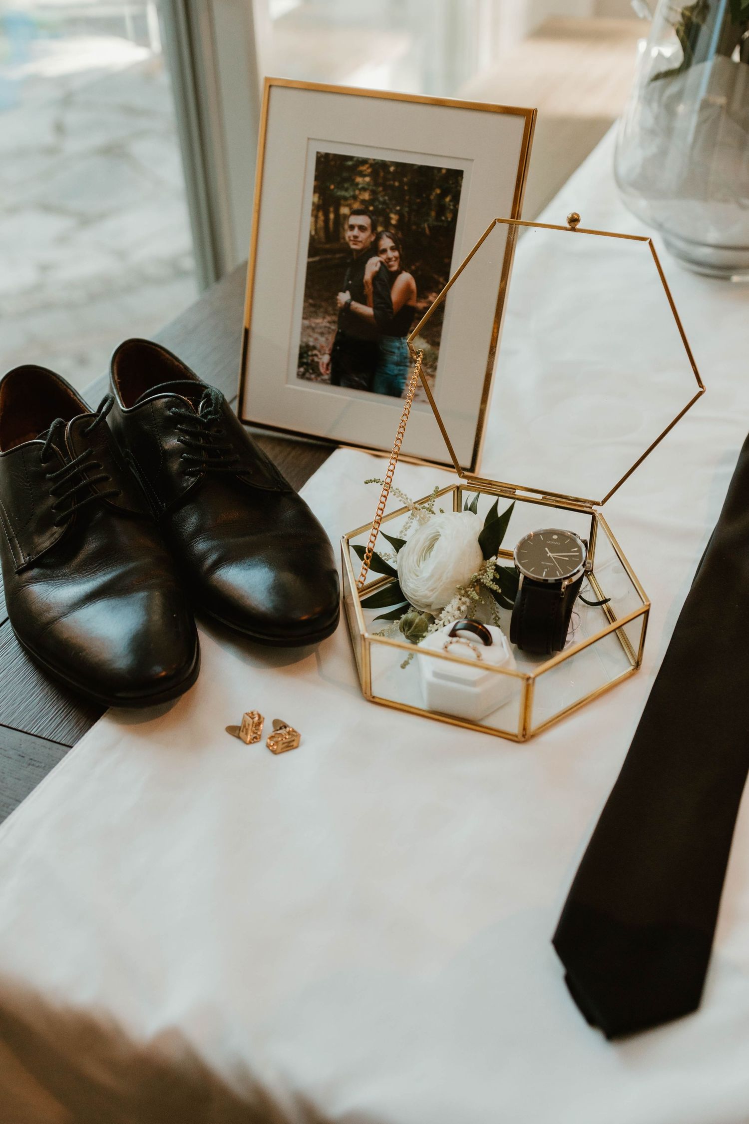Groom's wedding flat lay with black dress shoes, gold ring box, watch, cufflinks, tie and couple photo.