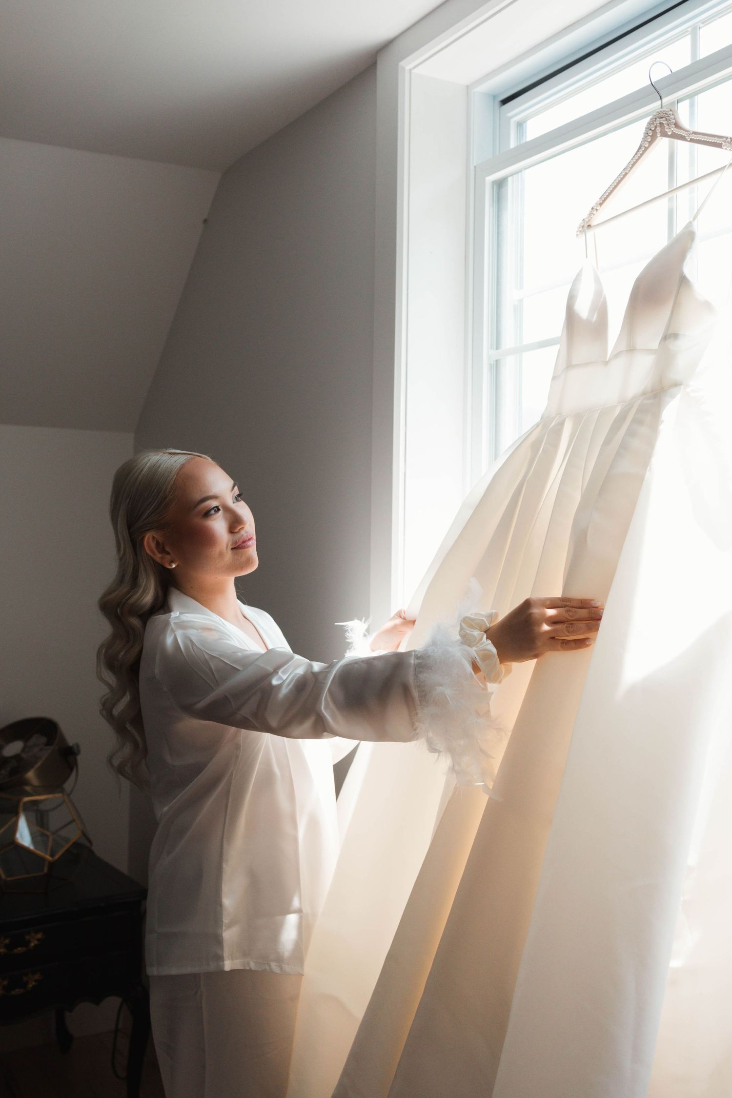 Blonde bride in white robe admiring wedding dress hanging by sunlit window.