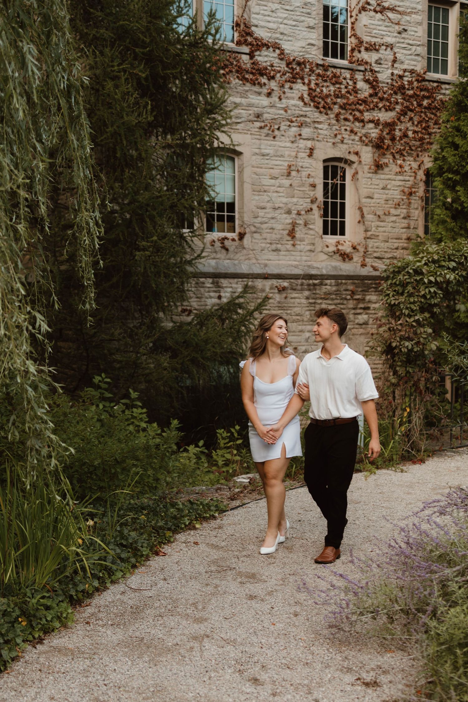 Engaged couple walking hand in hand on a garden path beside an ivy-covered stone building.