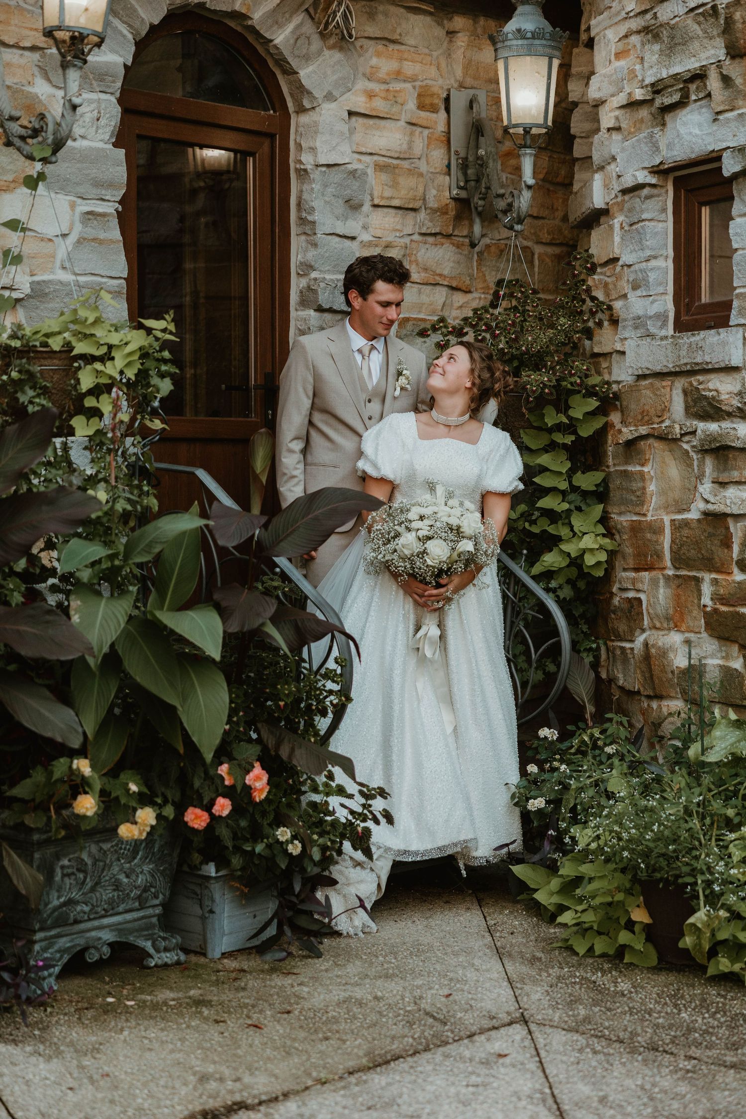 Bride and groom posing outside stone building surrounded by lush greenery.