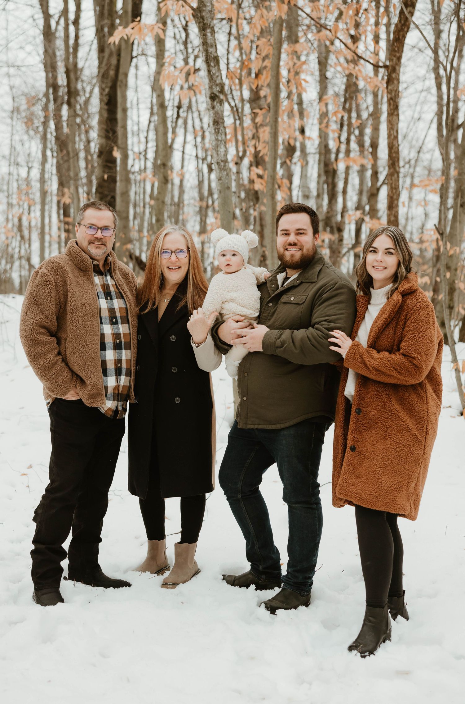 Family of five posing together in snowy winter woods with baby in white knit hat.